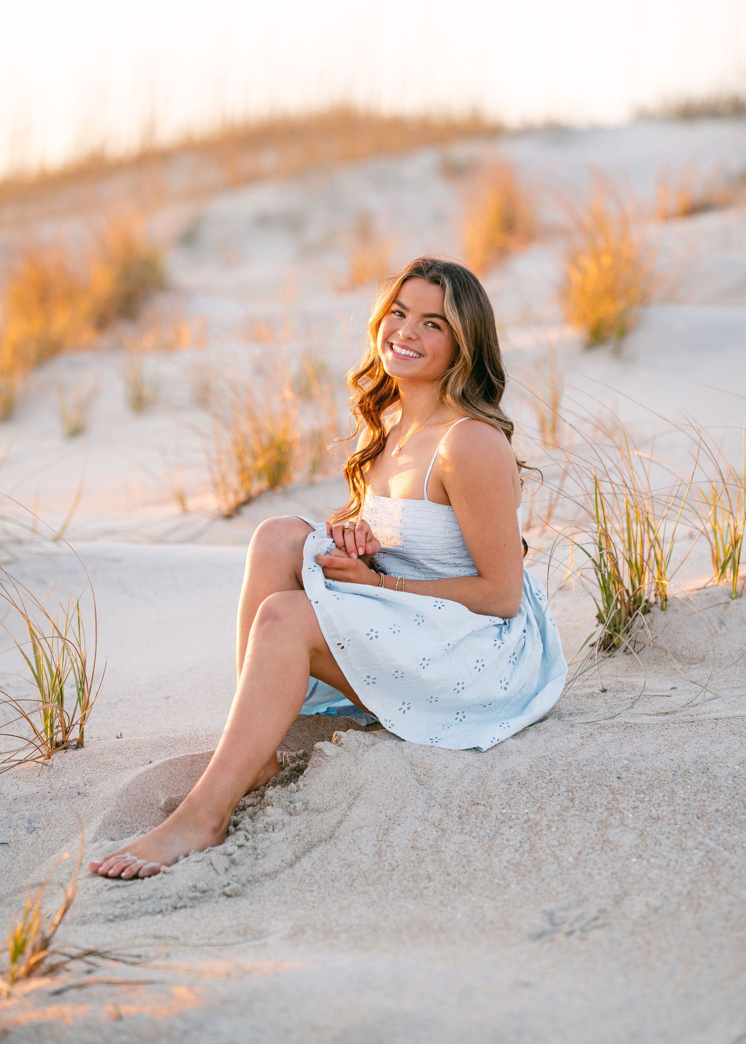 Smiling brunette woman in light blue sundress sitting on sandy beach dunes at golden sunset.