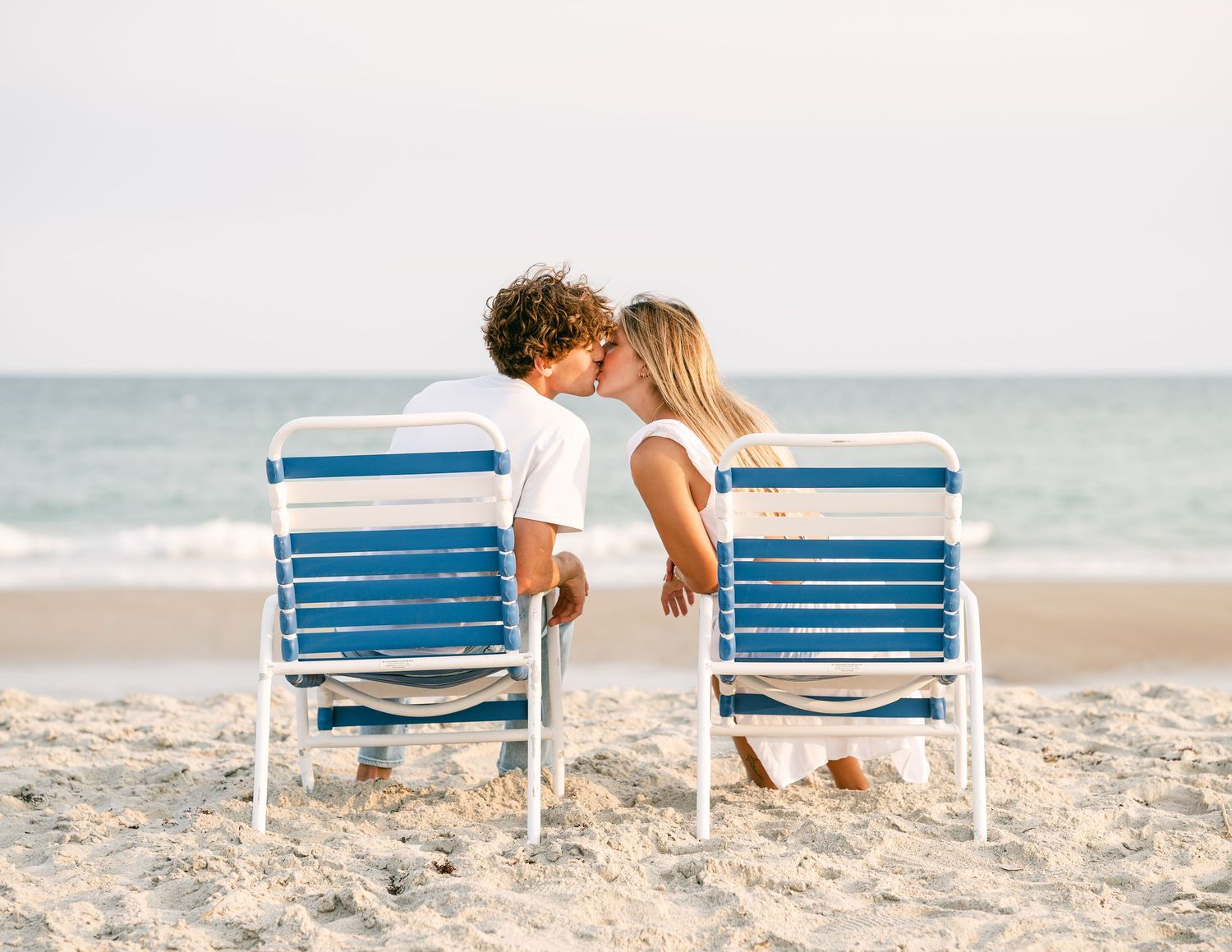 Couple sharing a romantic kiss while sitting in blue beach chairs on a sandy shore with ocean waves in the background.