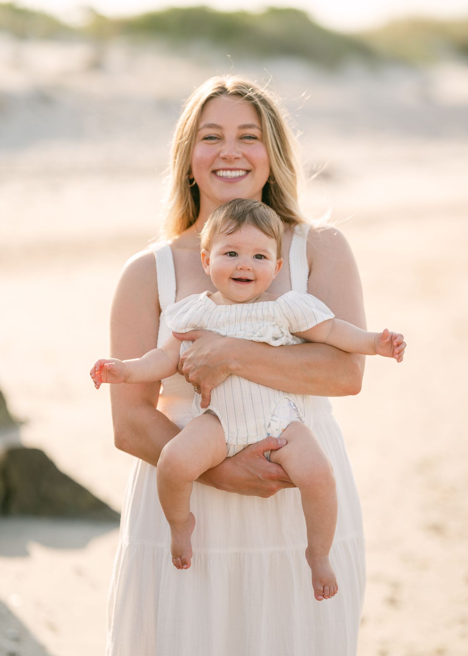 Smiling blonde mother in white dress holding happy laughing baby on sunny beach.