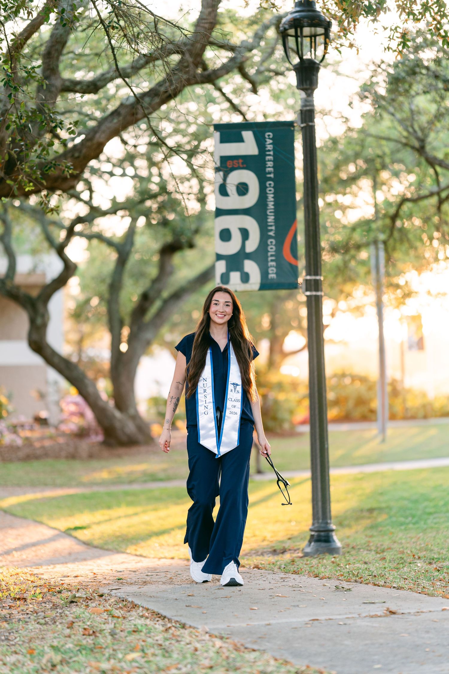 Smiling graduate in navy scrubs with stole walking past Carteret Community College 1963 banner.