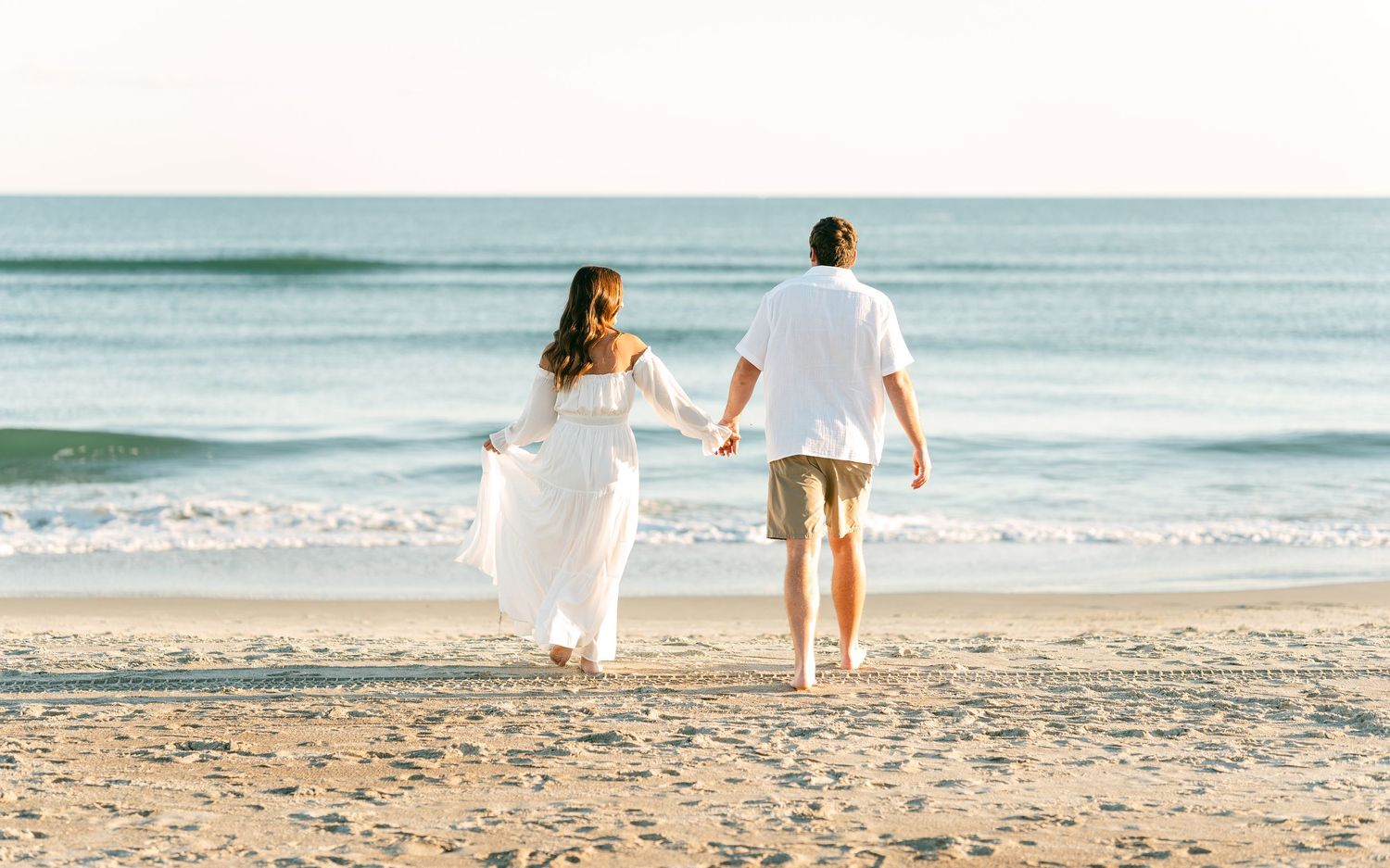 Newlywed couple holding hands walking along sandy beach shoreline in wedding attire.