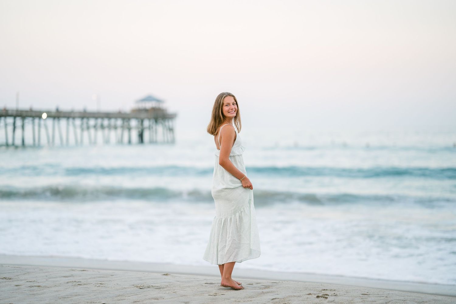 Young woman in white dress smiling on sandy beach at dusk with pier in background.