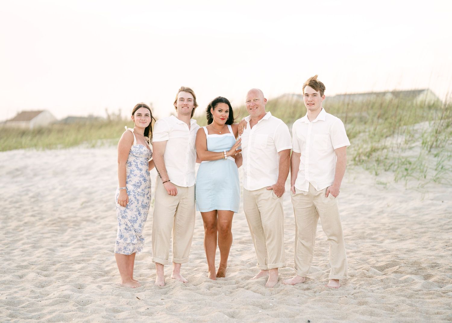 Family of five posing on a sandy beach with dunes and a house in the soft background light.