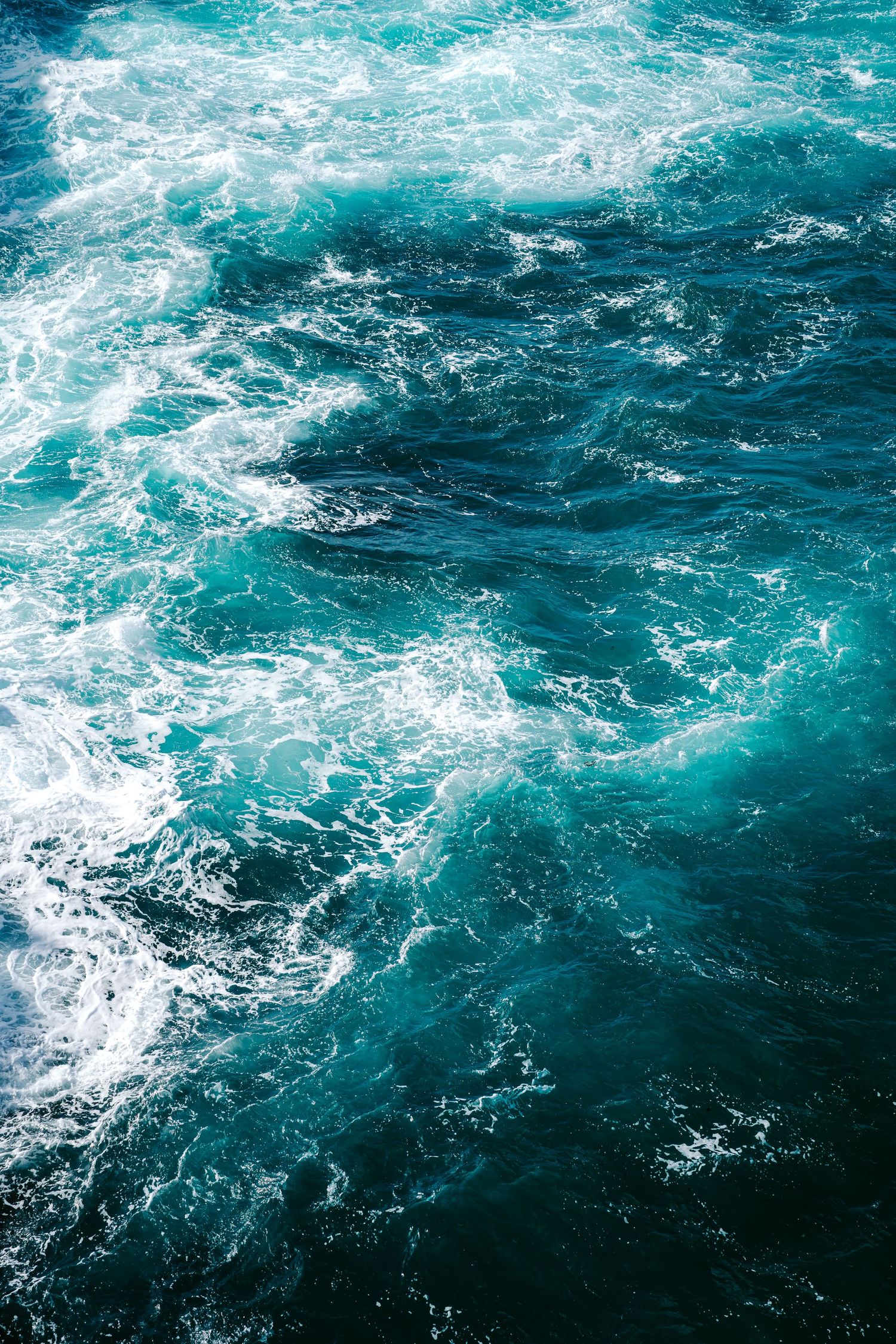 Aerial view of turbulent turquoise ocean waves with white sea foam swirling on the deep blue water surface.