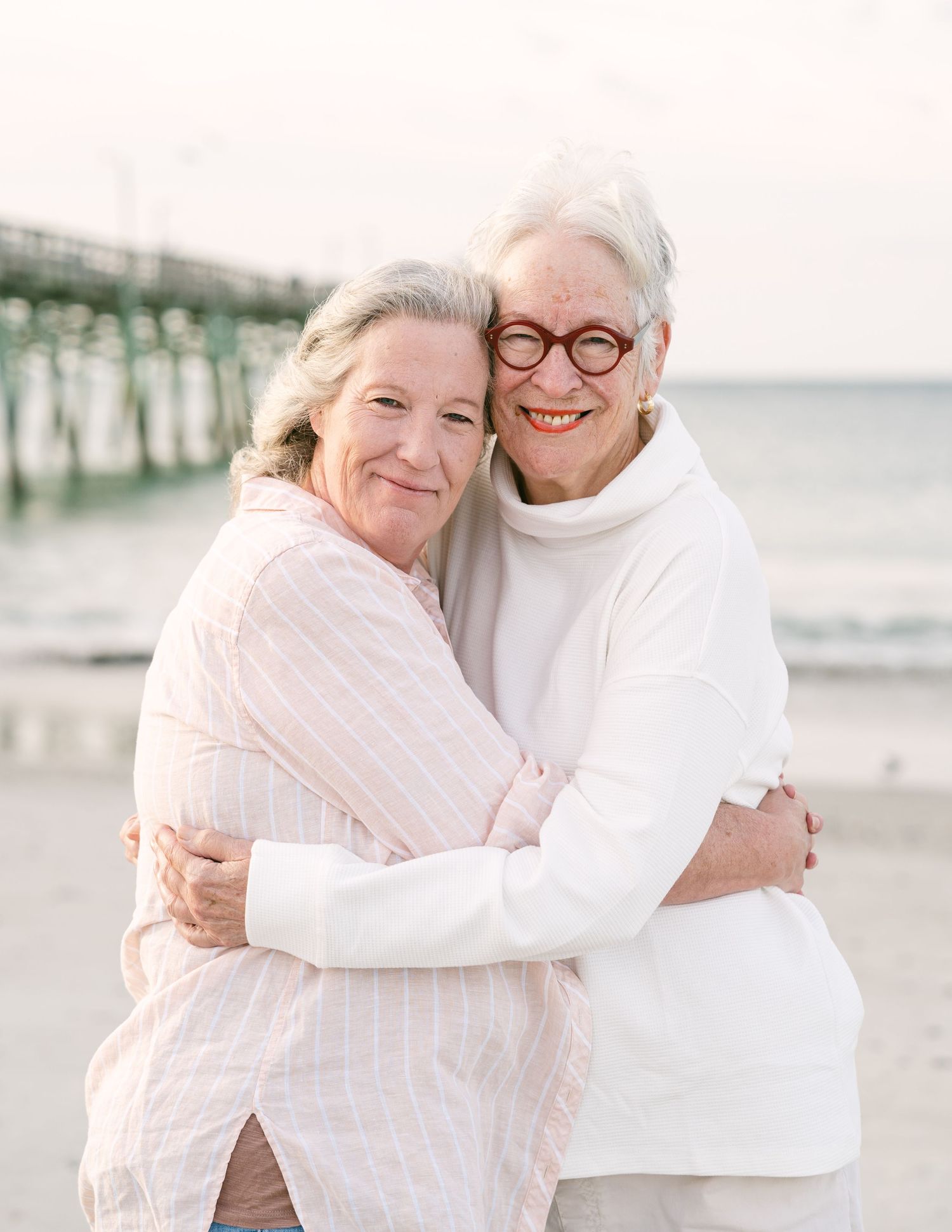 Two older women hugging warmly on beach near pier, smiling, wearing soft pink and white outfits.