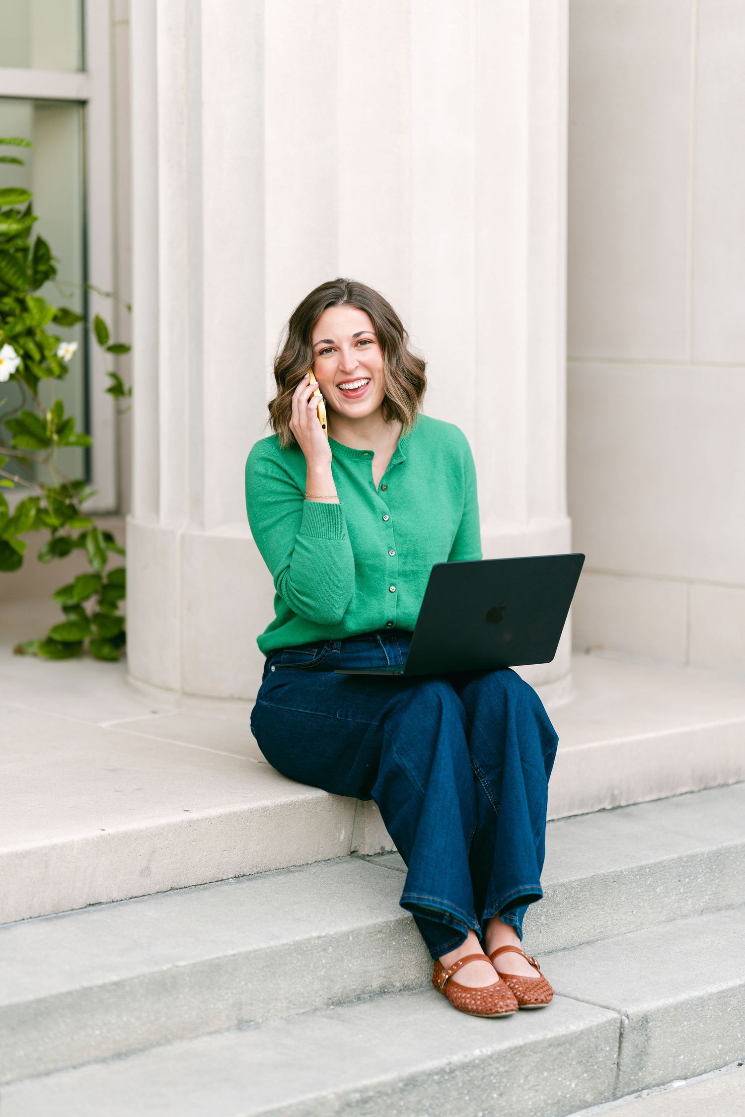 Smiling woman in green blouse sitting on steps with laptop, talking on phone outdoors.