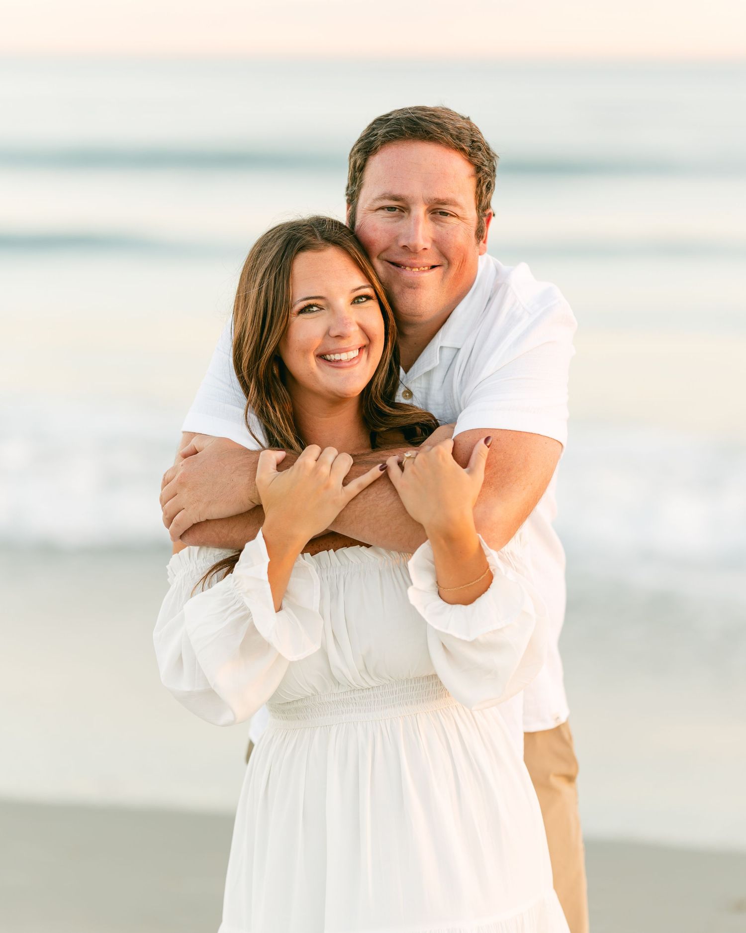 Happy couple embracing on a beach at sunset, both dressed in white outfits.