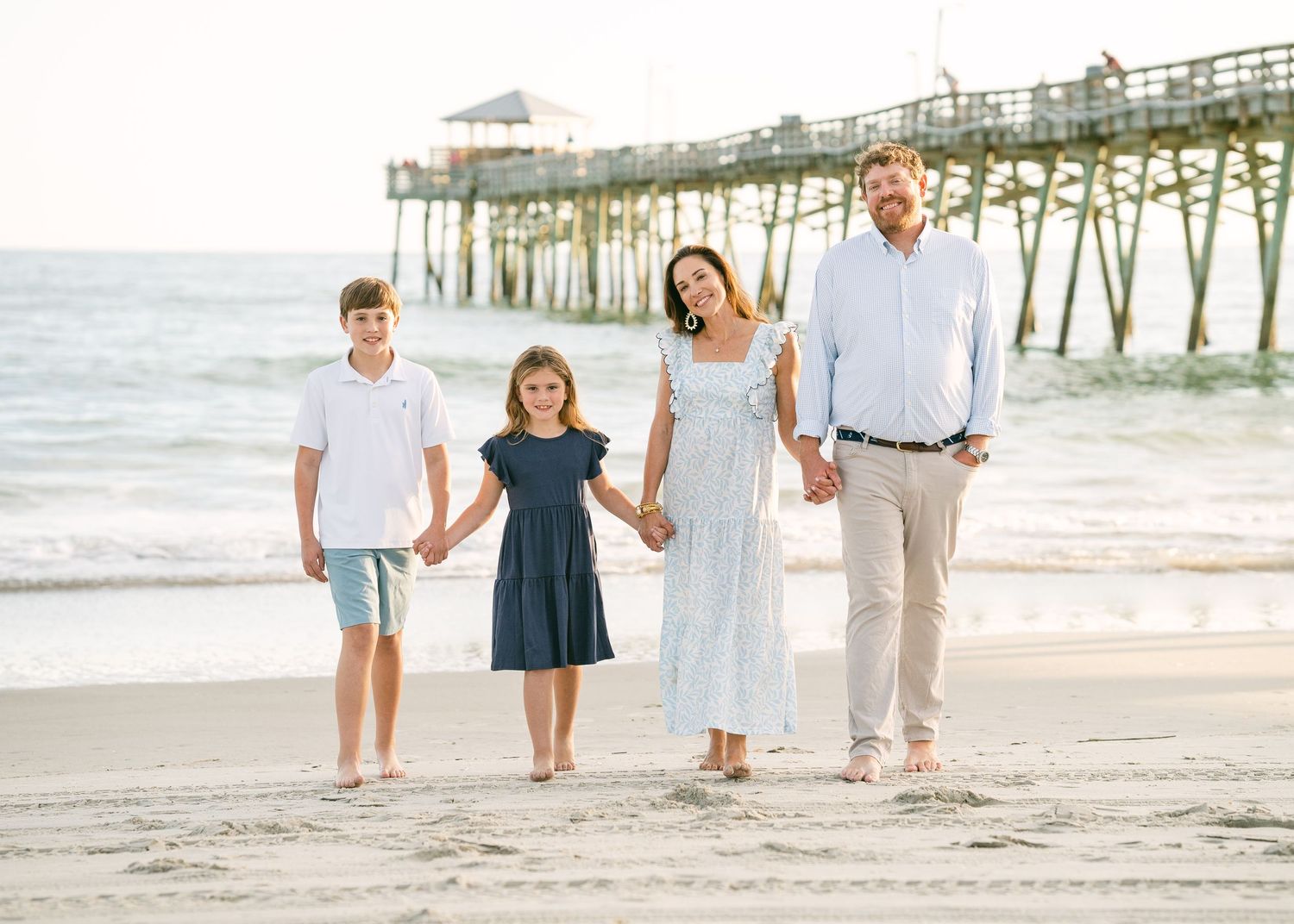 Smiling family of four posing together on sandy beach with ocean pier in background.