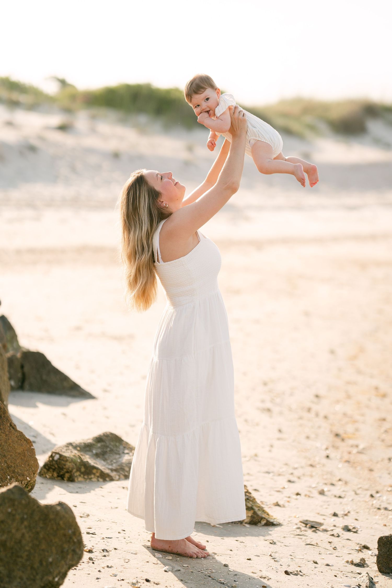 Mother in white dress lifting laughing baby overhead on sunny sandy beach at sunset.