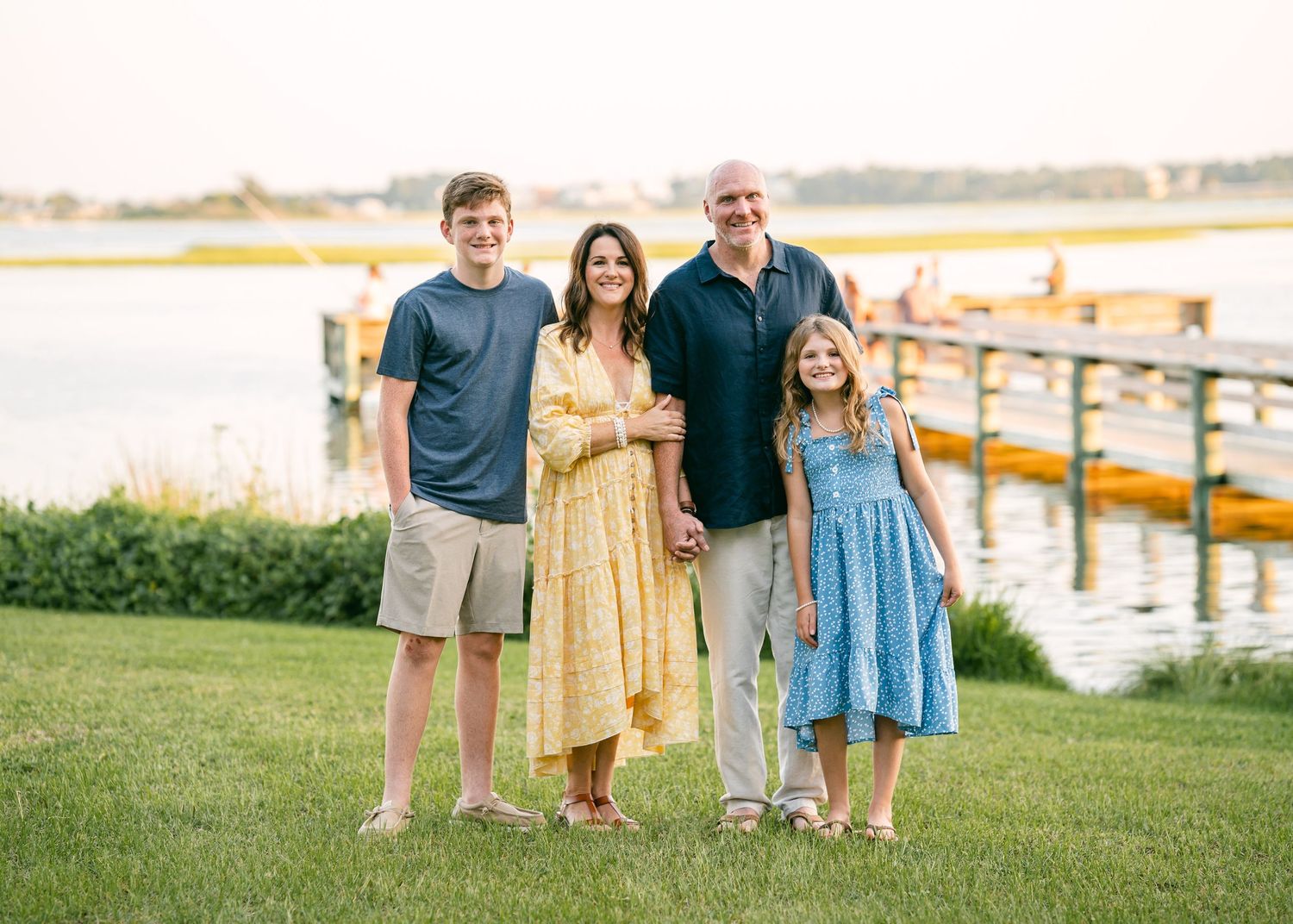 Family of four smiling together on green lawn with waterfront dock in background.