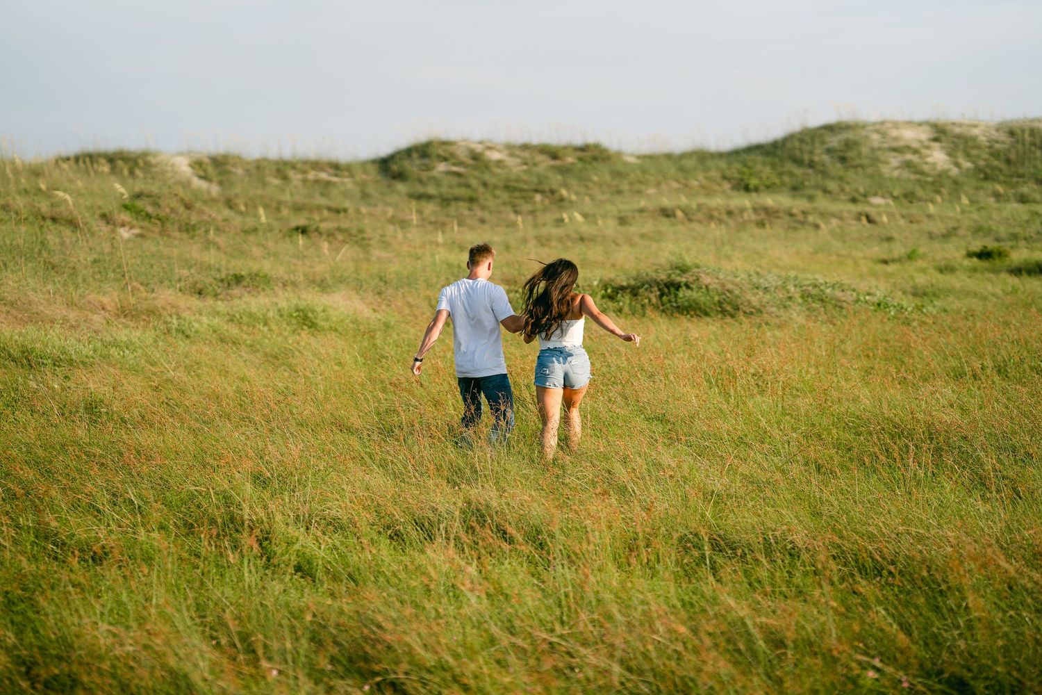 Couple holding hands running through tall green grass fields with sand dunes behind them.