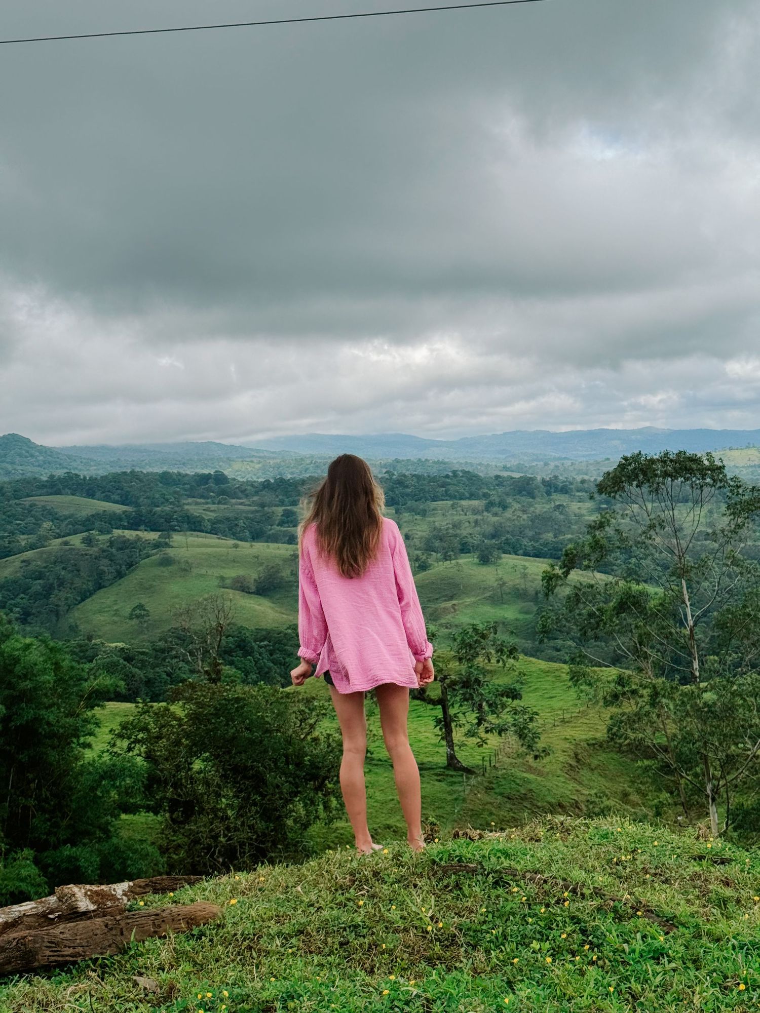 Woman in pink oversized shirt stands on hilltop overlooking lush green tropical valley landscape.