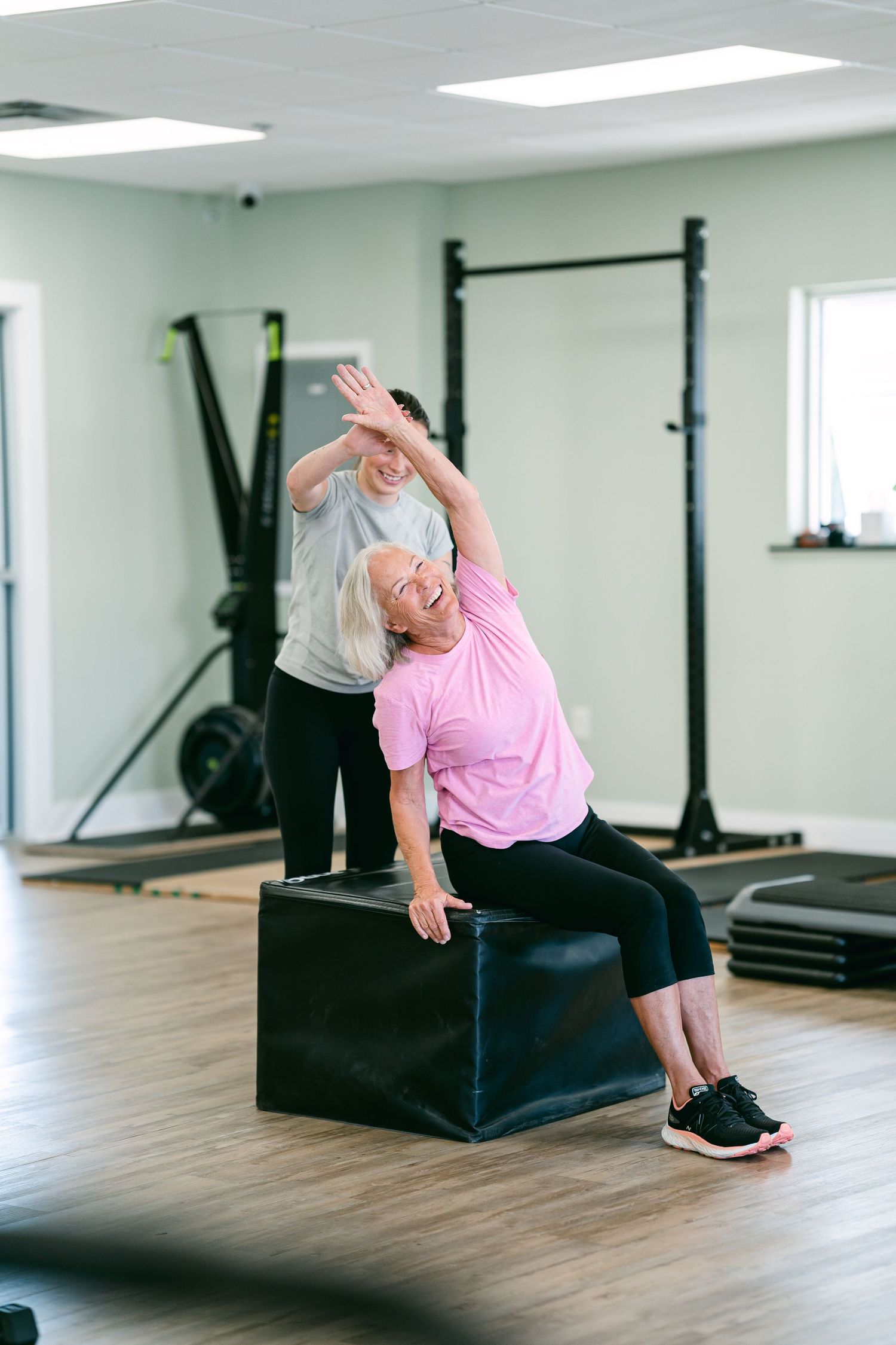 Personal trainer assists senior woman in pink with lateral stretch exercise on plyo box in bright gym.