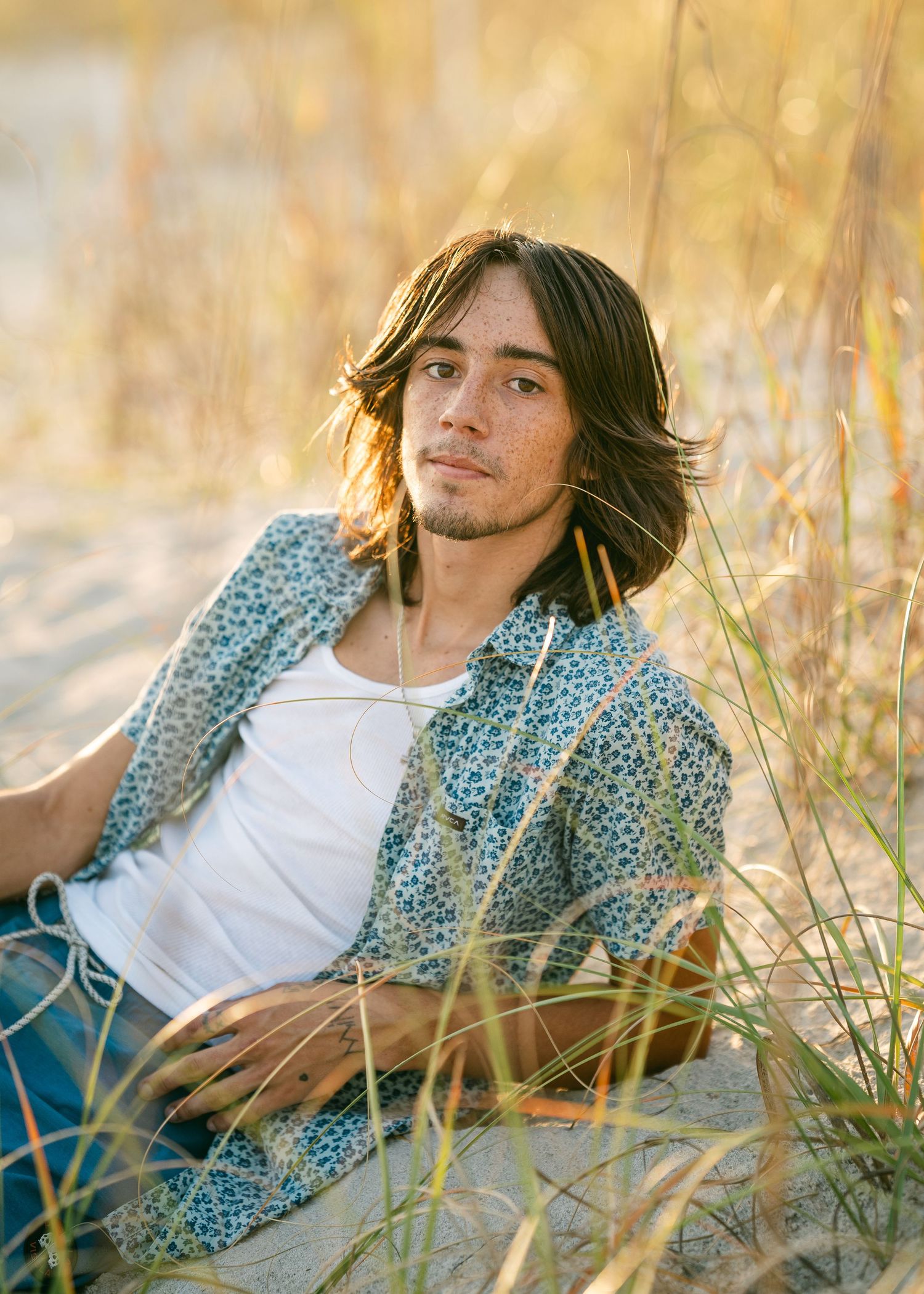 Young man with long hair sitting on sandy beach dunes surrounded by tall grass.