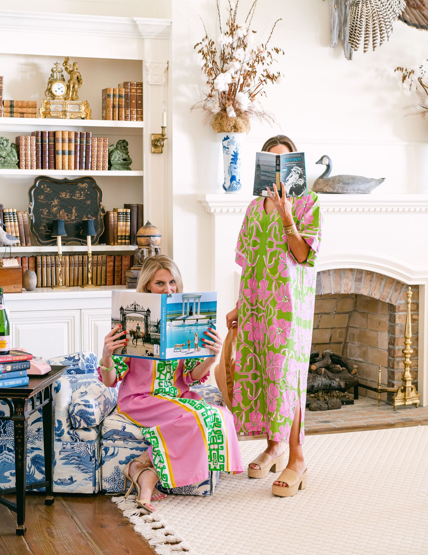 Two women in colorful Lilly Pulitzer dresses reading books in a chic living room with fireplace.