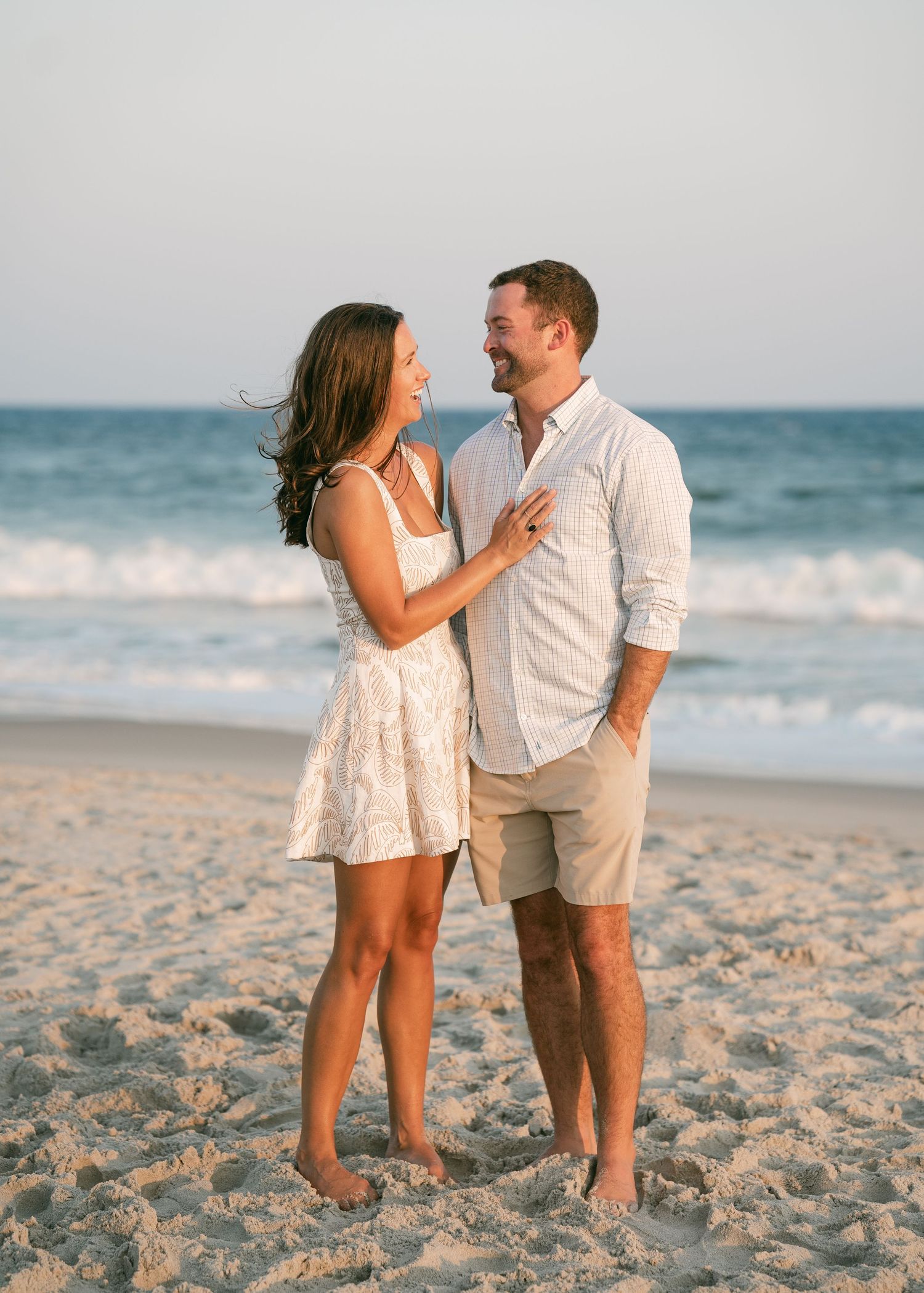 Couple smiling at each other on a beach with ocean waves behind them.