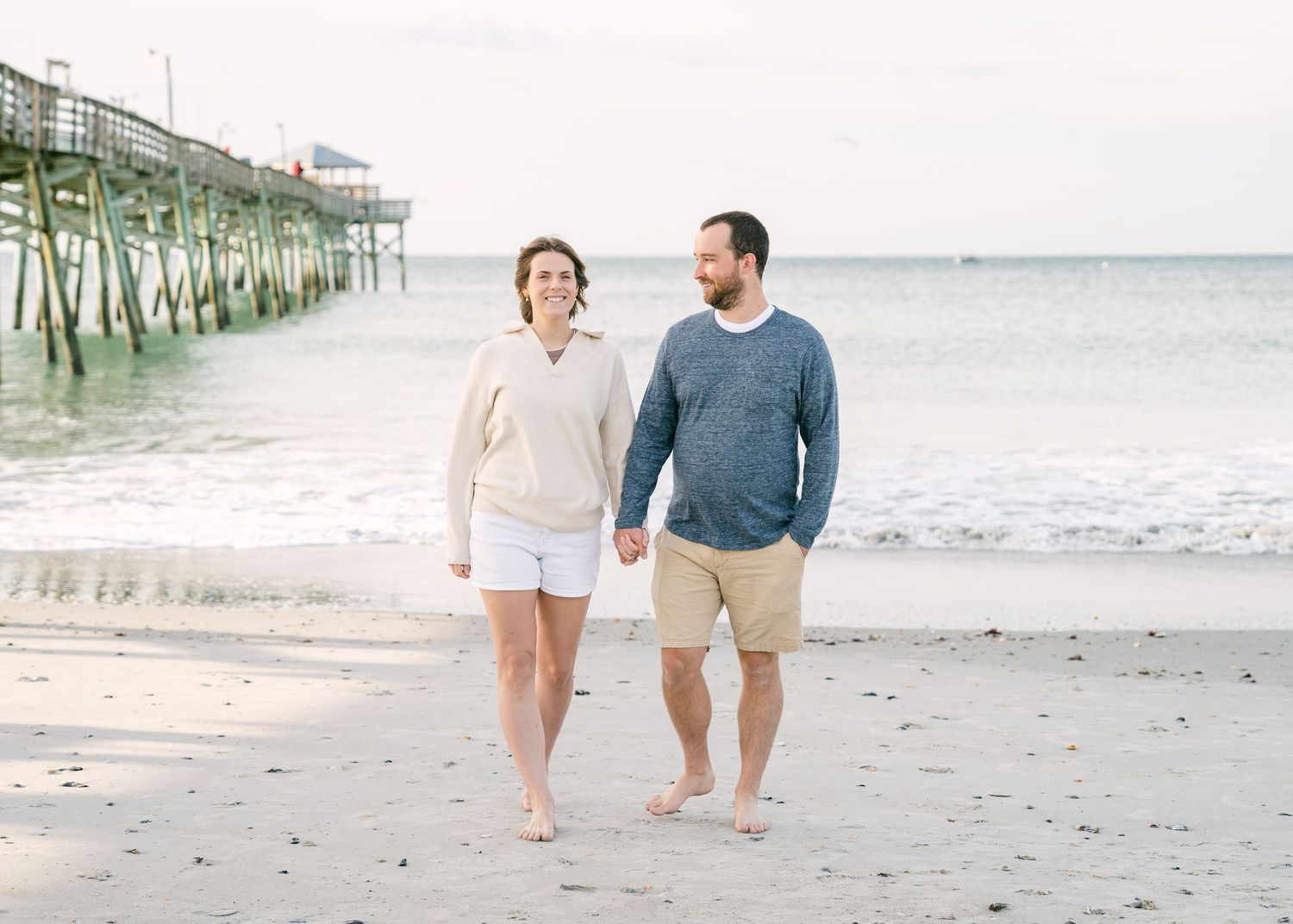 Couple holding hands walking barefoot along shoreline near beach pier, dressed in casual coastal attire.