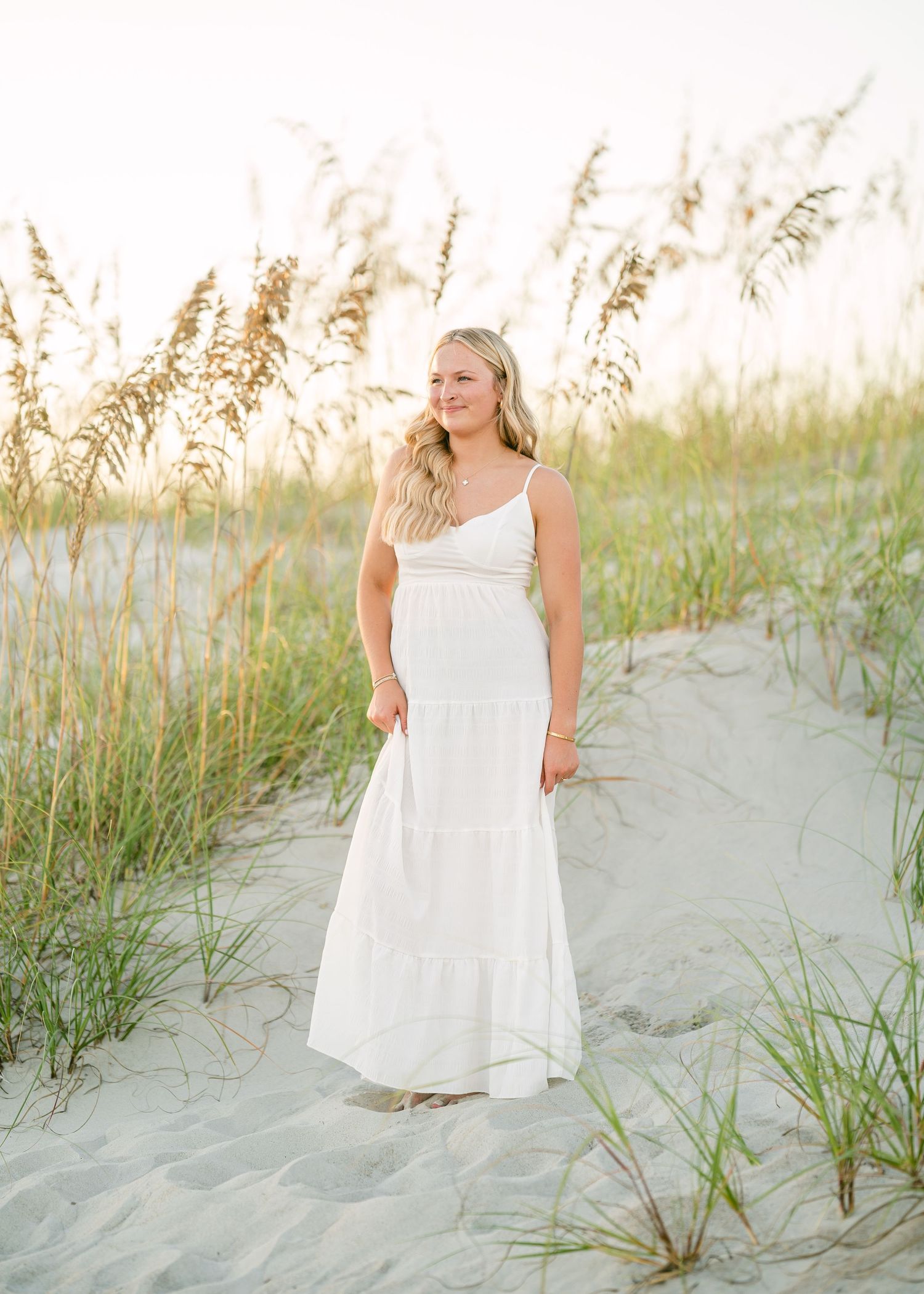 Blonde woman in white tiered maxi dress standing on sandy beach path surrounded by sea oats at sunset.