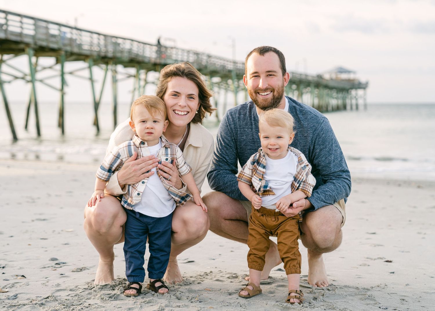 Young family with twin toddler boys crouching on beach near pier, smiling in coordinated fall outfits.