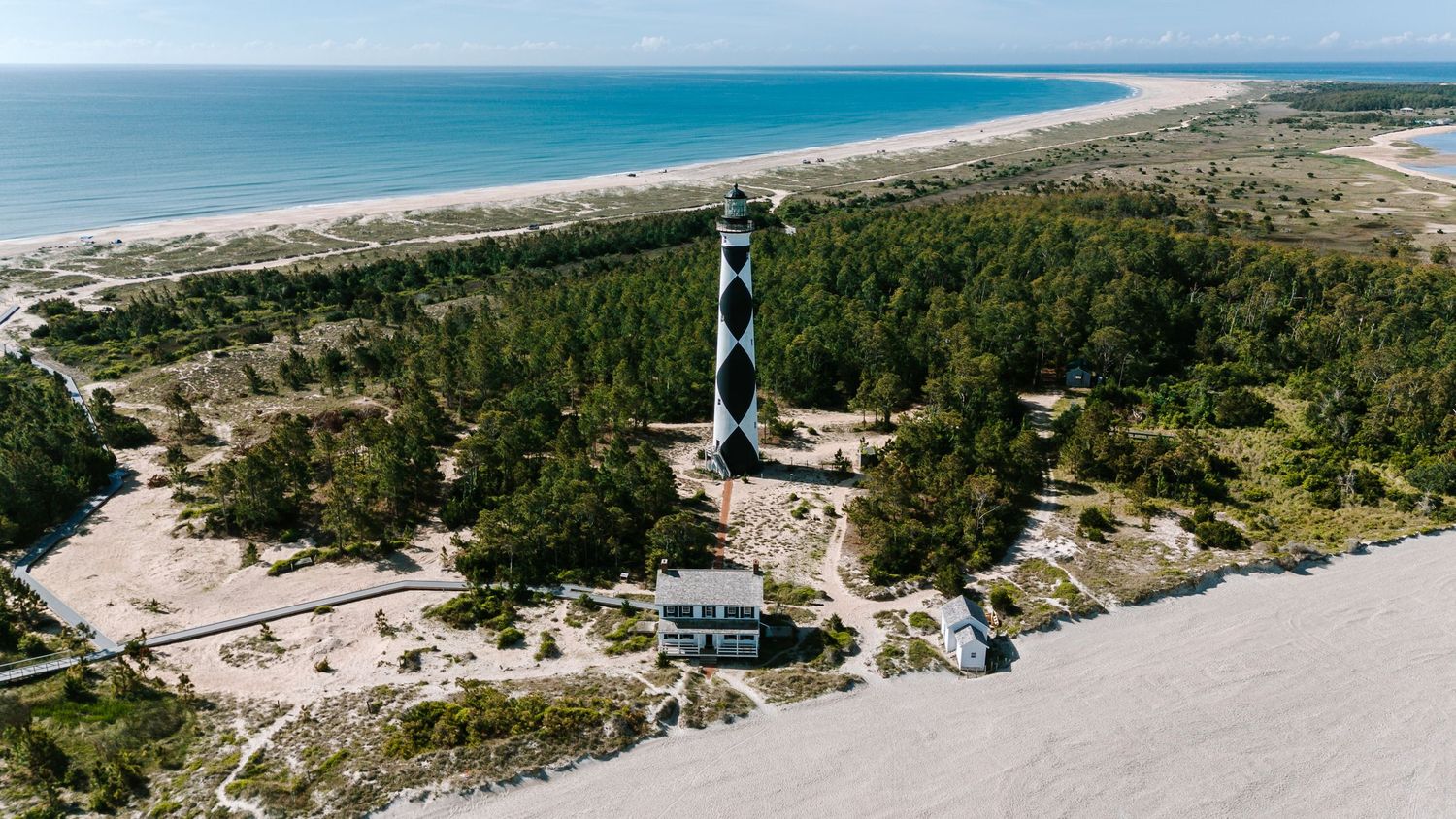 Aerial view of Cape Lookout Lighthouse surrounded by trees near a sandy beach coastline.