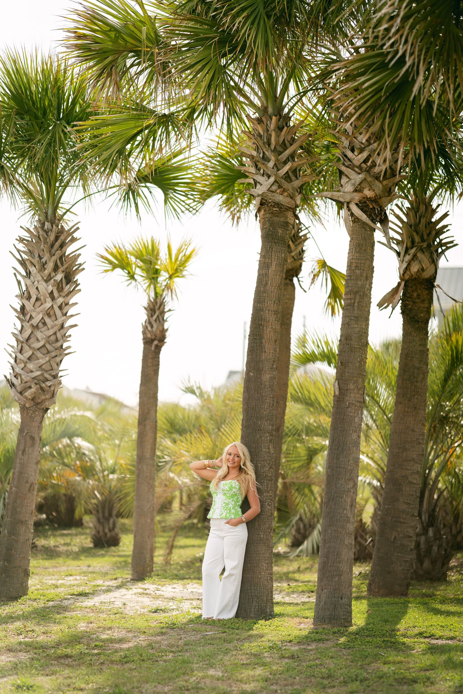 Blonde woman in green top and white pants leaning against palm tree in tropical setting.
