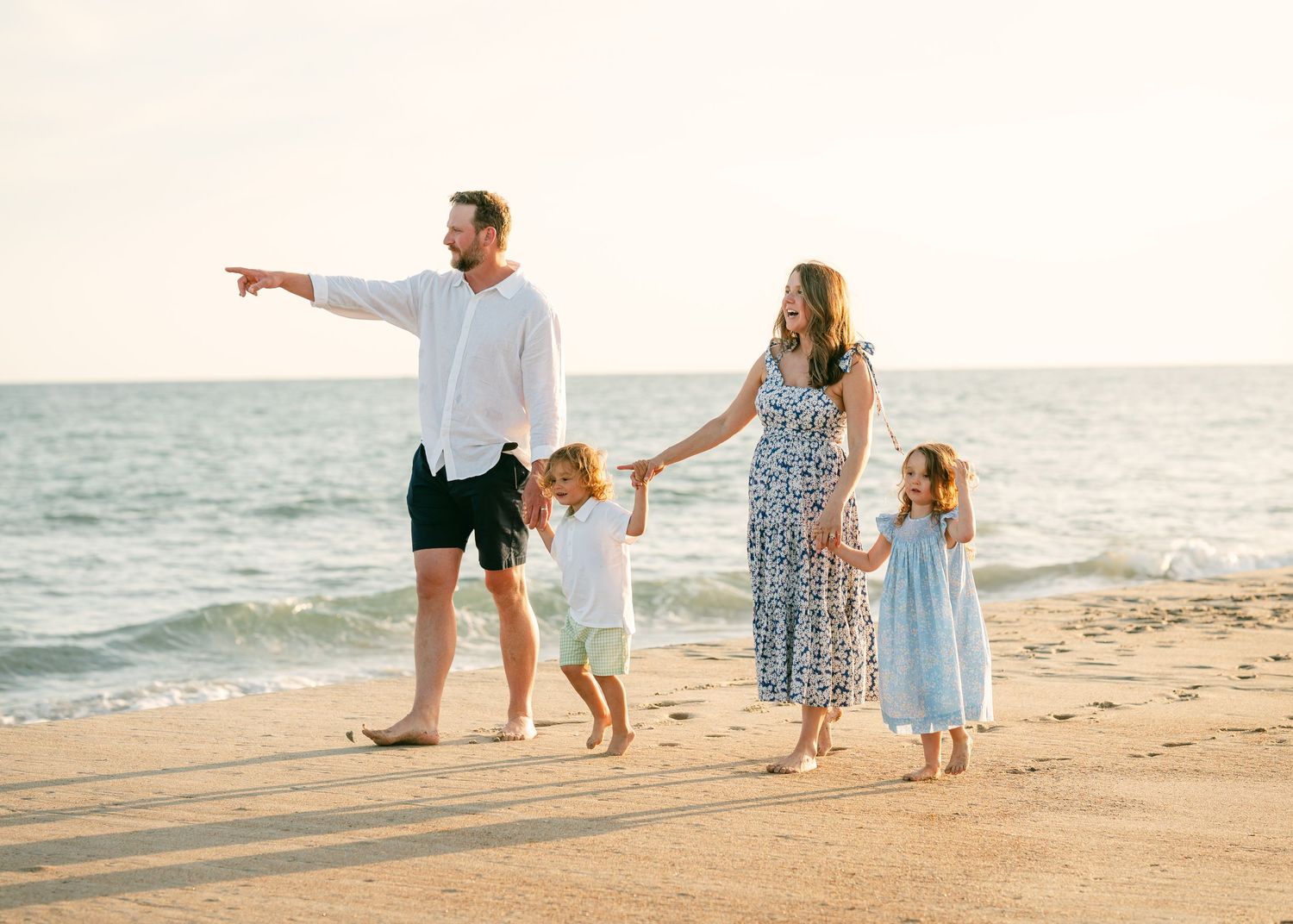 Family of four walking on sandy beach at sunset, father pointing at ocean.