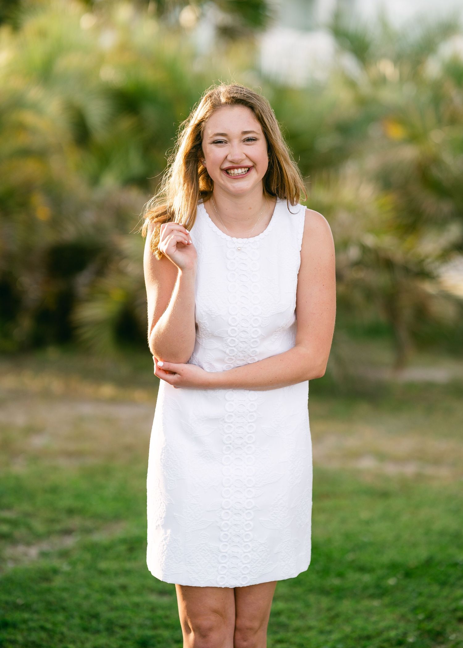 Laughing teen girl in white sleeveless dress posing outdoors with palm trees for senior photo.