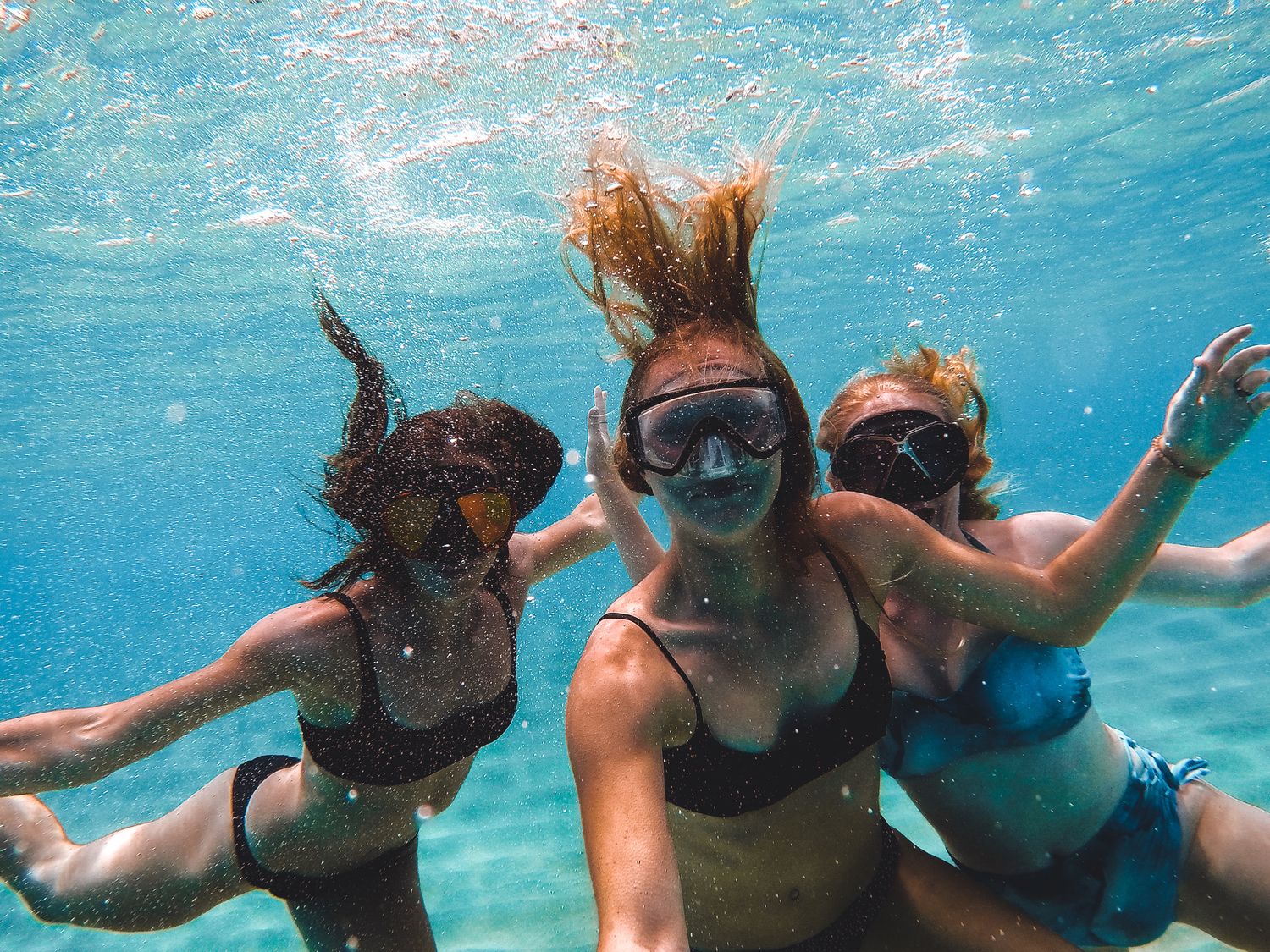 Three women swimming underwater in clear blue water, one wearing snorkel goggles.