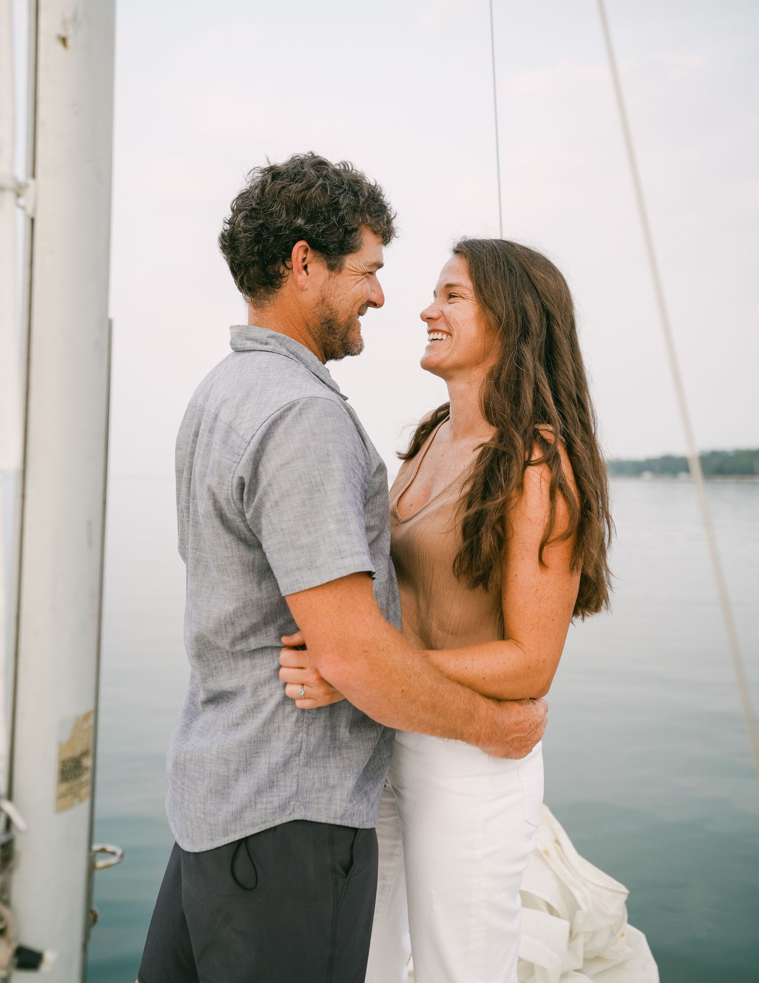 Happy couple laughing and embracing on a sailboat on calm water during engagement photos.