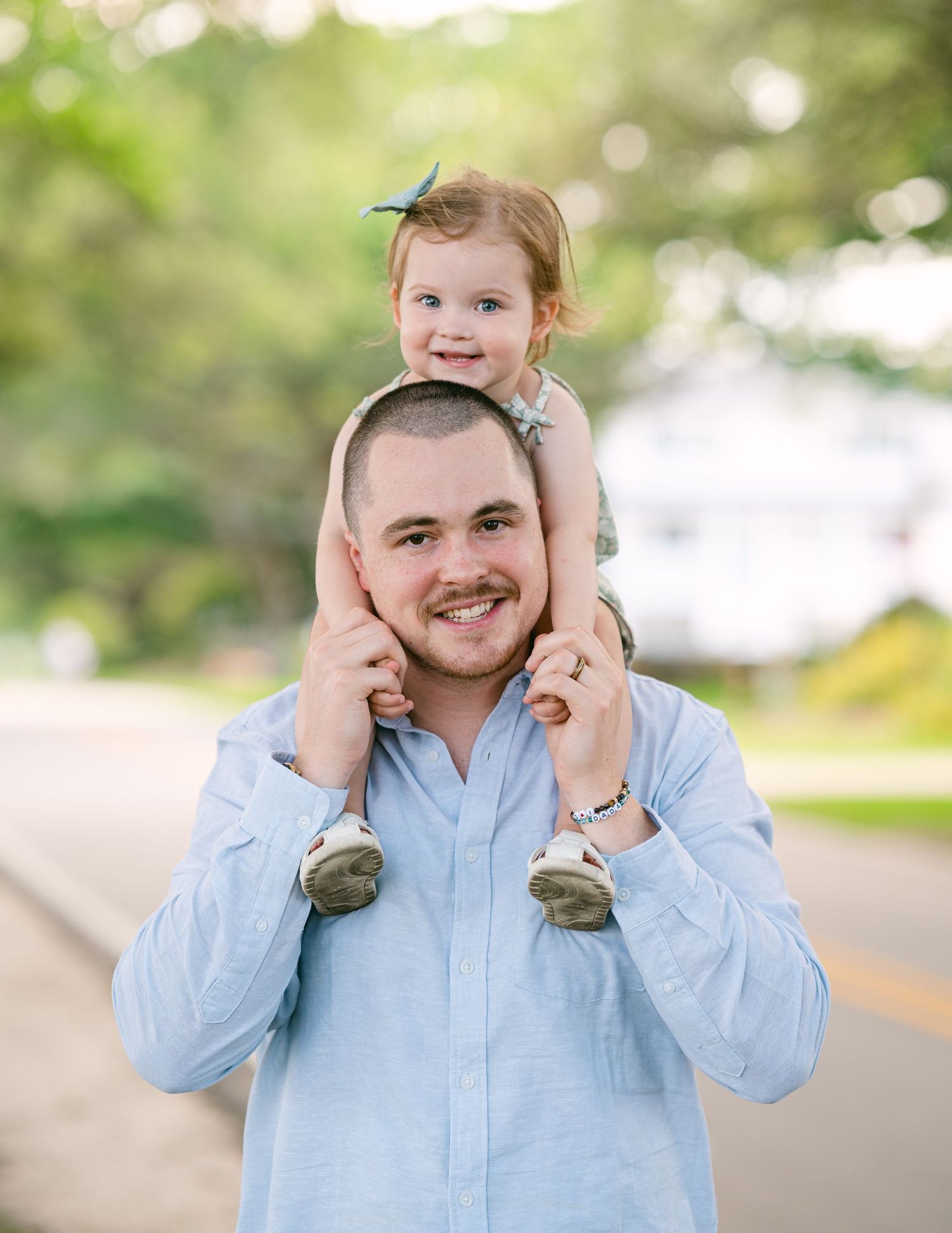 Smiling father carrying happy toddler girl on his shoulders on a sunny tree-lined street.