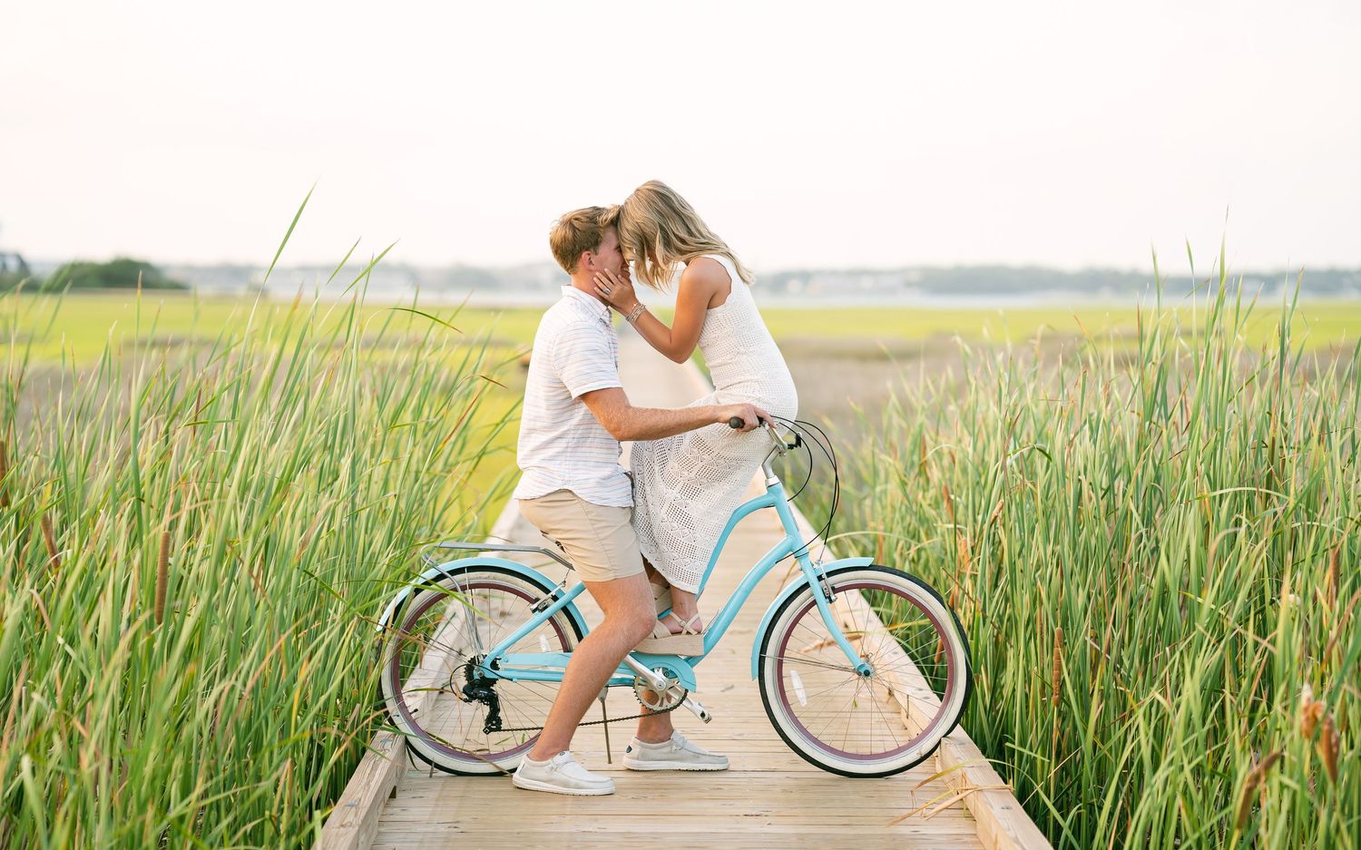 Couple sharing a kiss on a teal beach cruiser bike on a wooden dock in marsh.