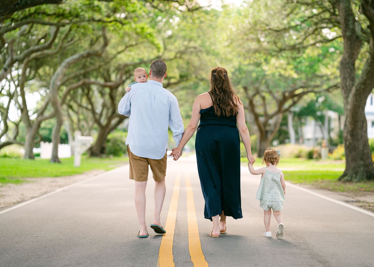 Family of four walking away down a sunny tree-lined road, holding hands together.