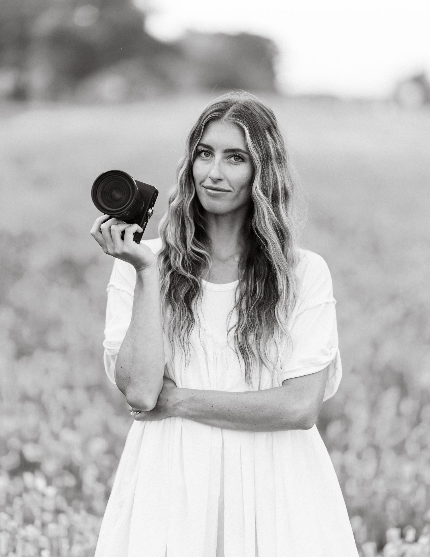 Black and white portrait of a woman in a white dress holding a camera in a flower field.