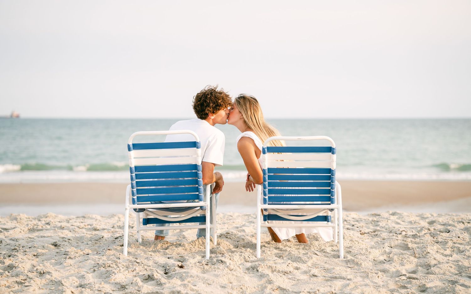 Young couple sharing a kiss while sitting in beach chairs facing the ocean.