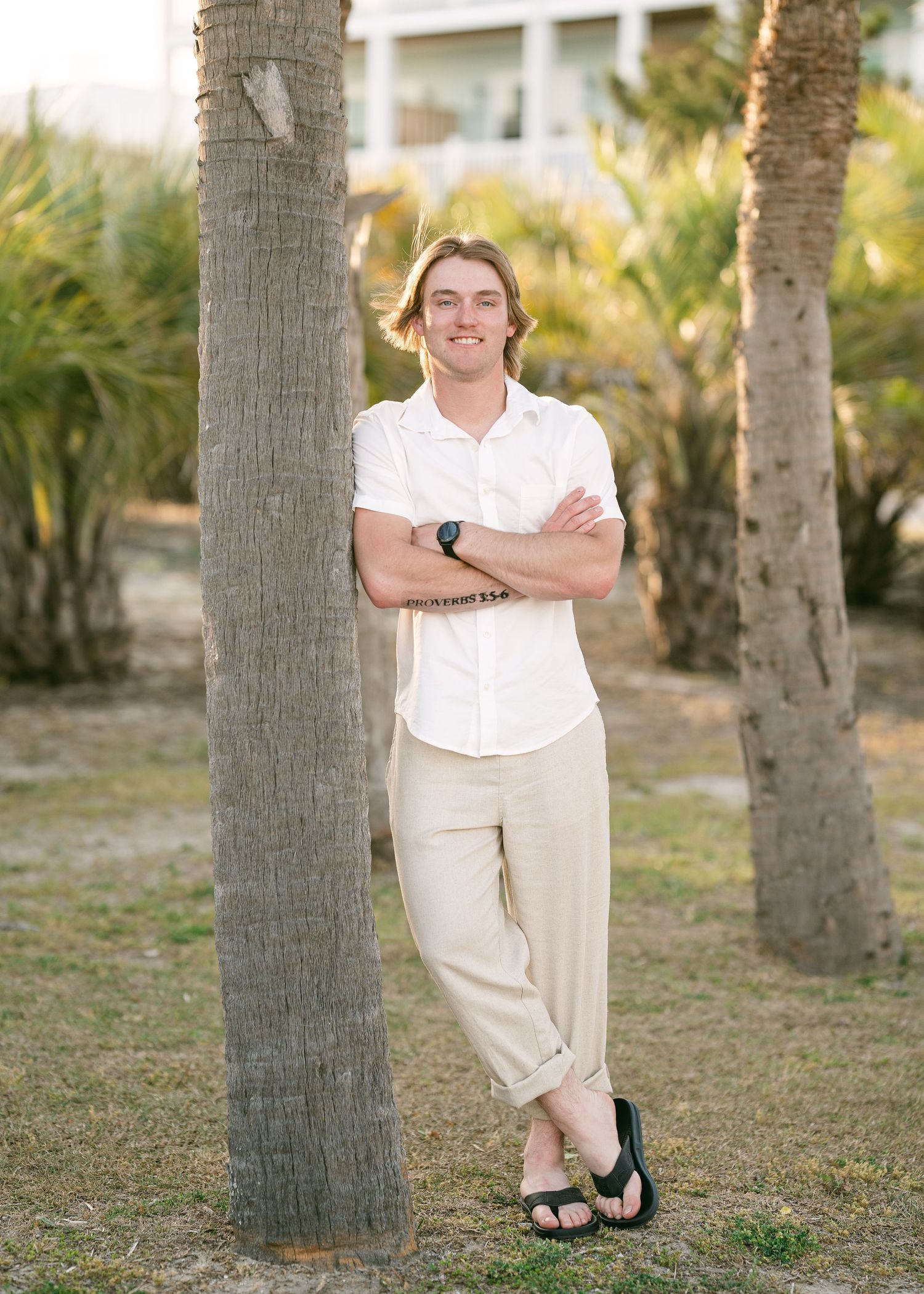 Teenage boy in white shirt and khaki pants leaning against a palm tree in a sunny outdoor setting.