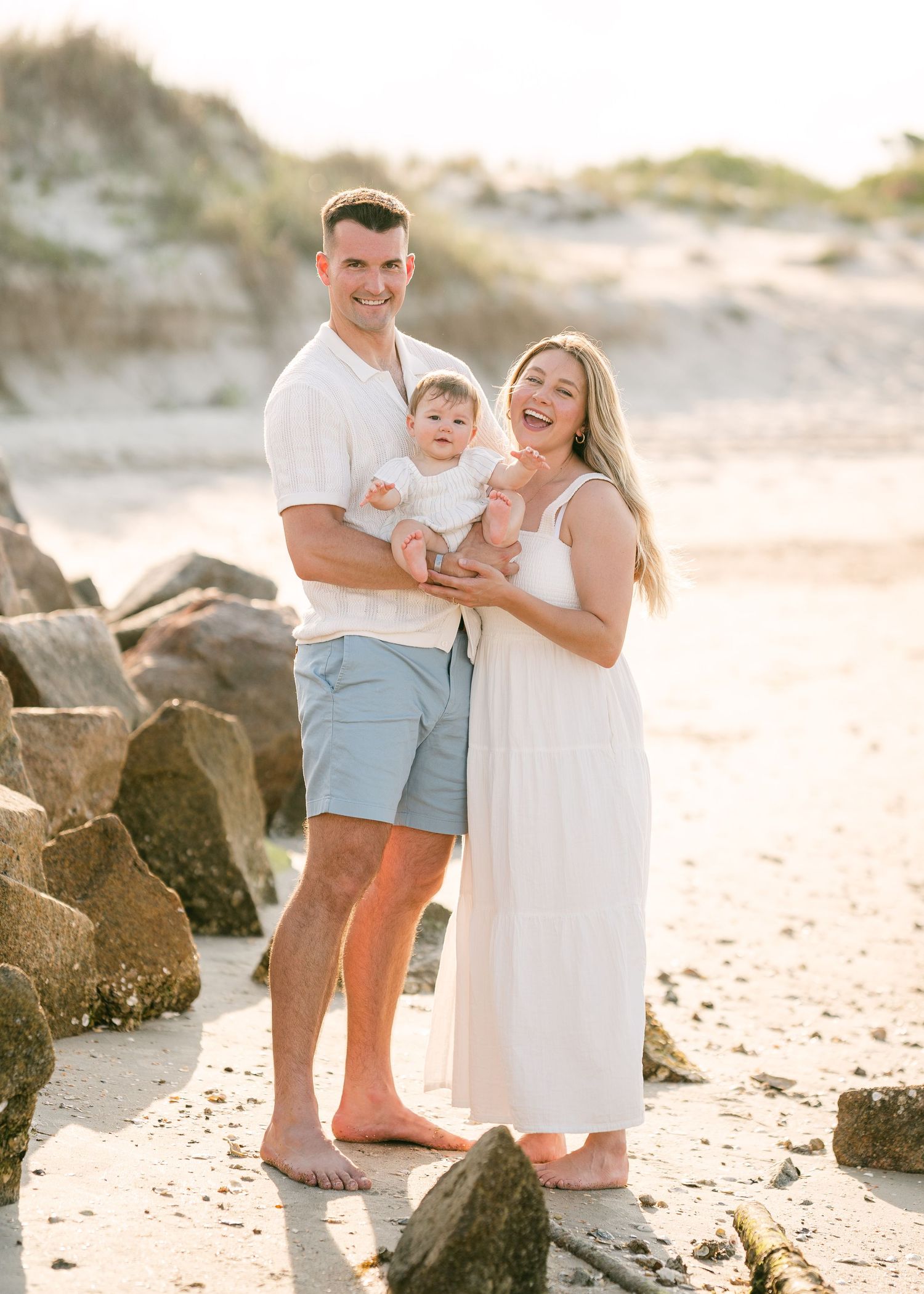 Smiling family of three standing near rocks on beach, baby in white outfit at golden hour.