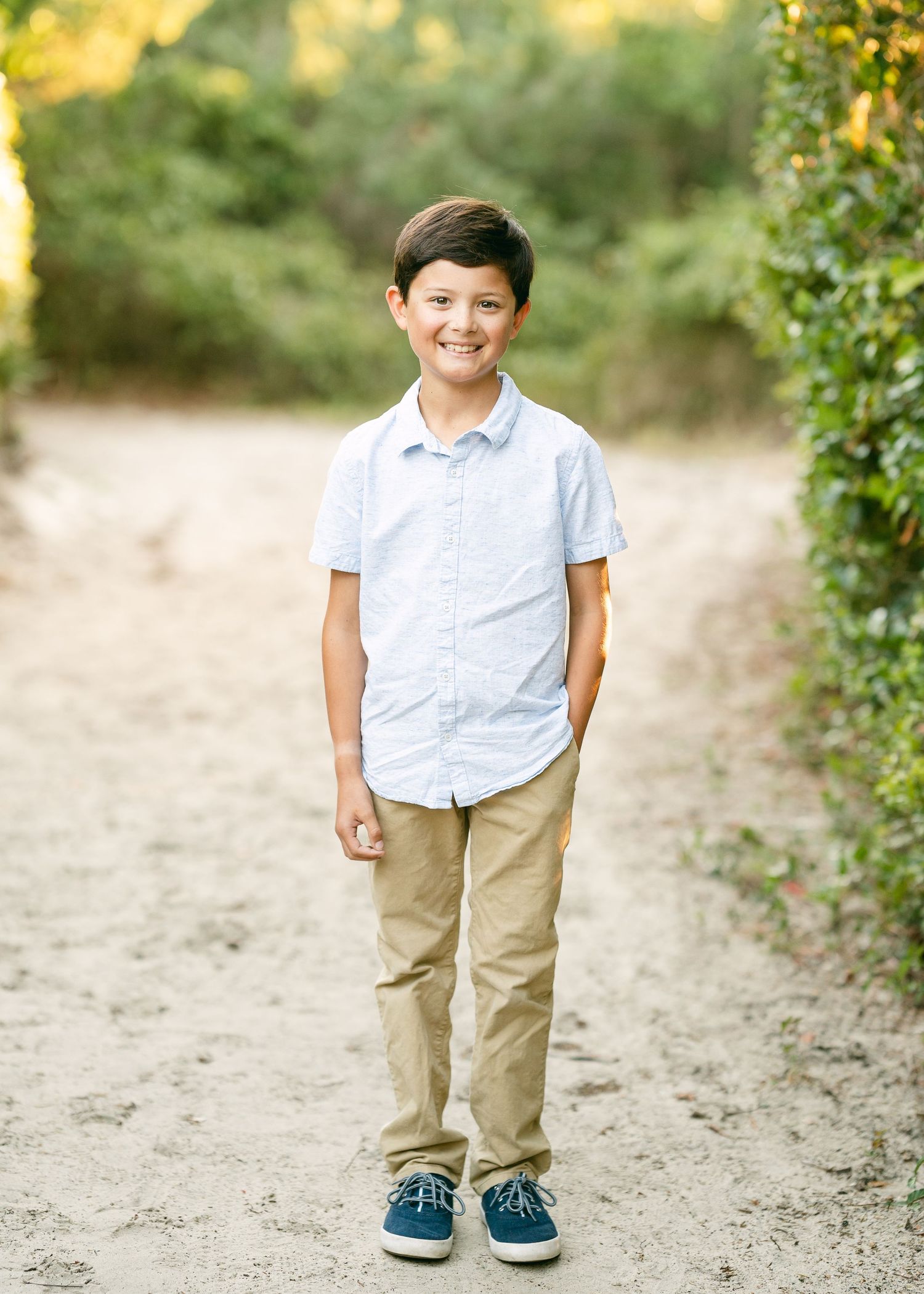 Smiling young boy in a light blue shirt and khaki pants standing on a sandy path.
