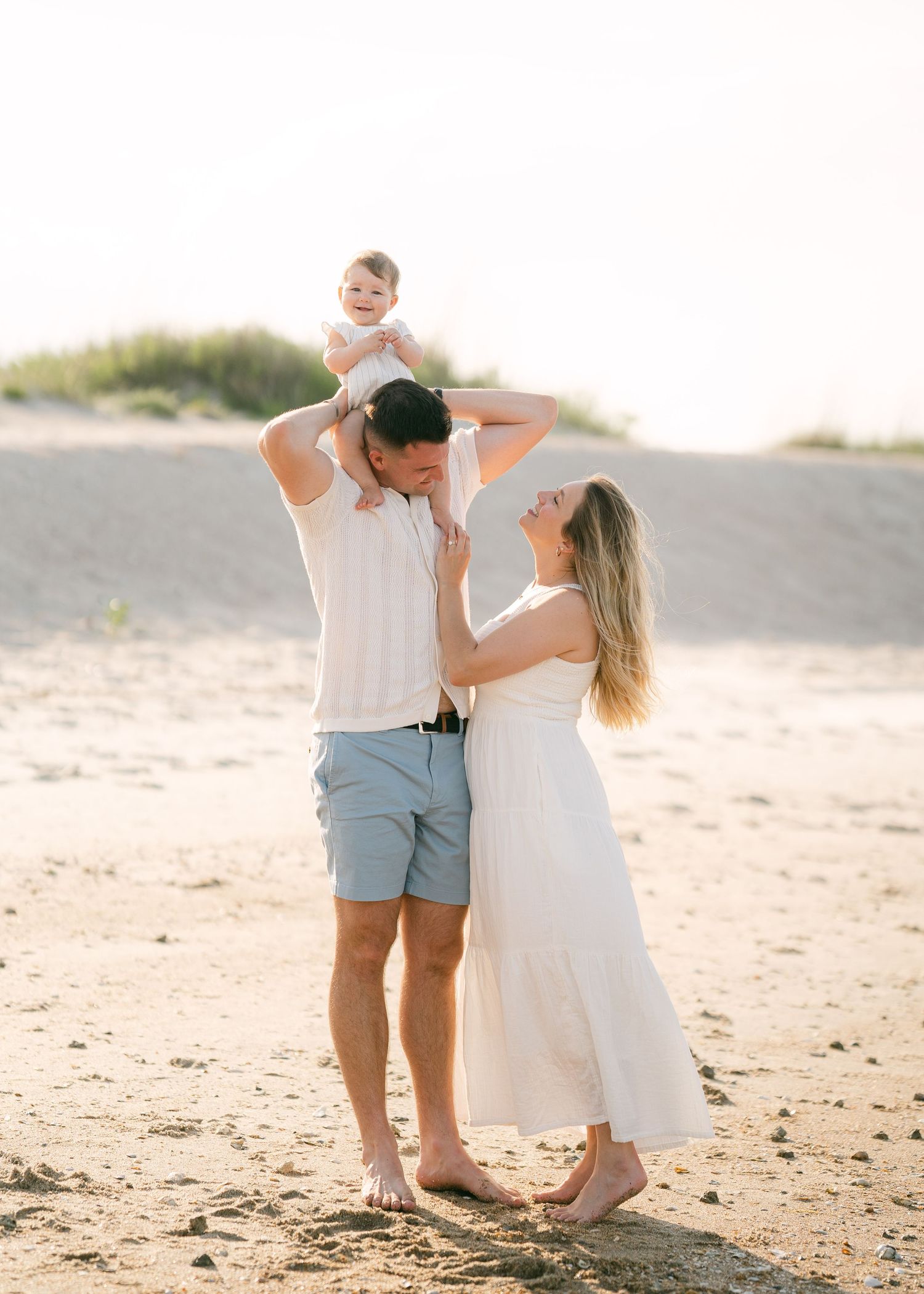 Happy family lifting baby overhead on sandy beach, mother in white dress laughing at sunset.