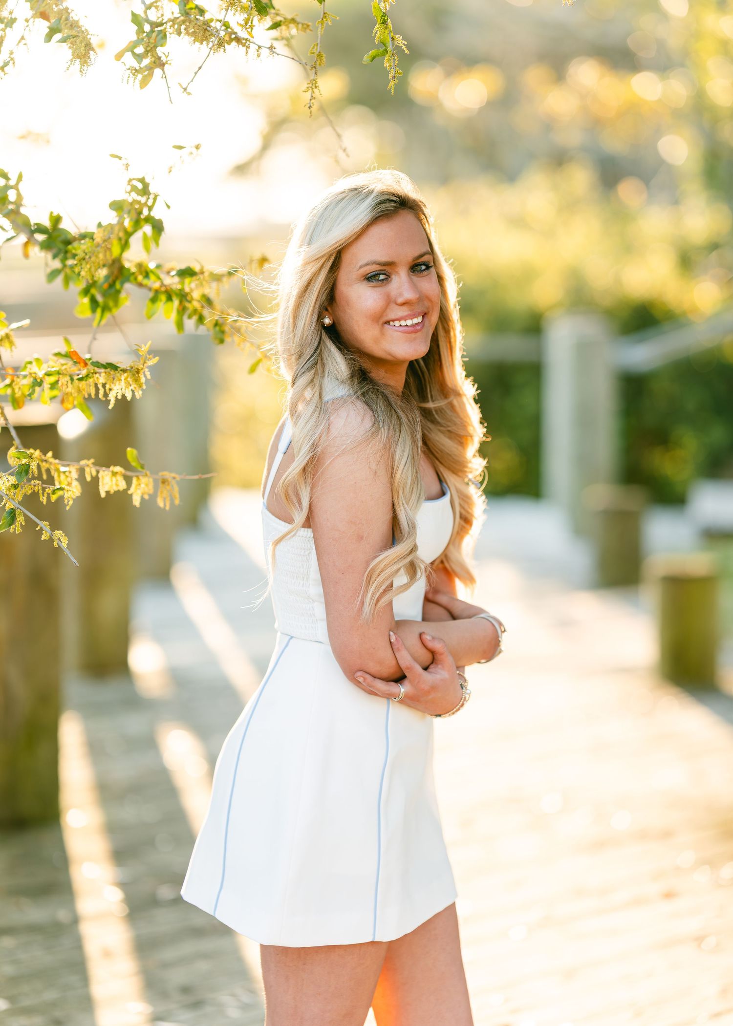 Smiling blonde woman in white tennis dress posing on sunny wooden boardwalk surrounded by trees.