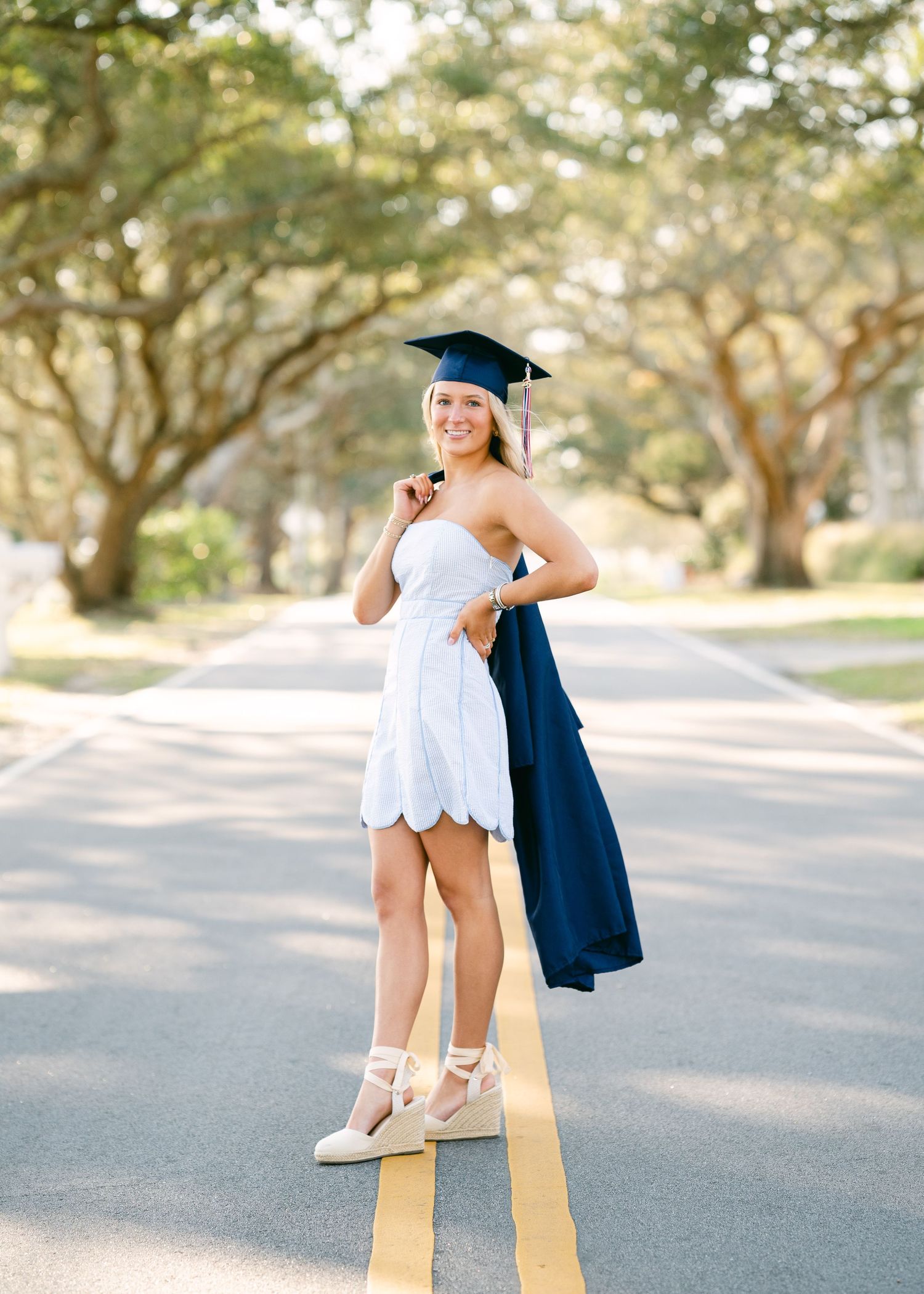 Blonde graduate in white dress and navy cap and gown posing on tree-lined road.