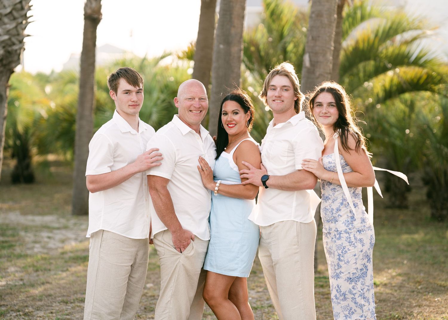 Happy family of five posing together outdoors among palm trees in coordinated beige and white outfits.