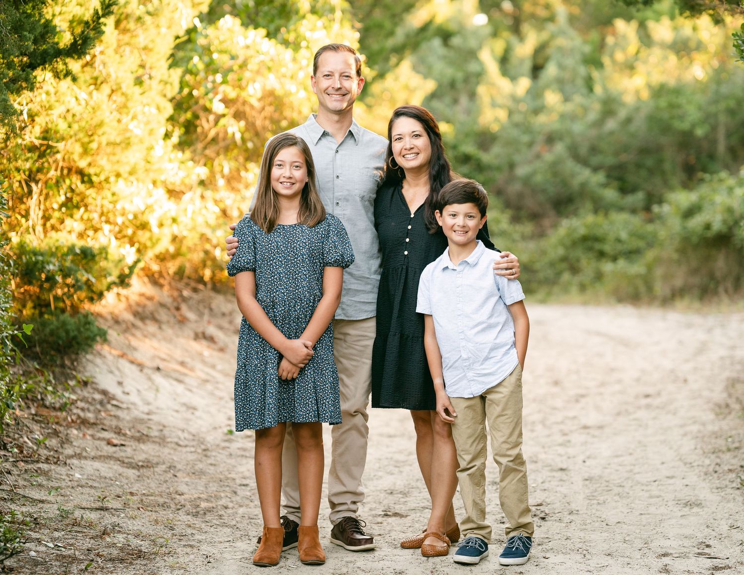 Happy family of four posing on a sandy trail surrounded by lush green foliage.