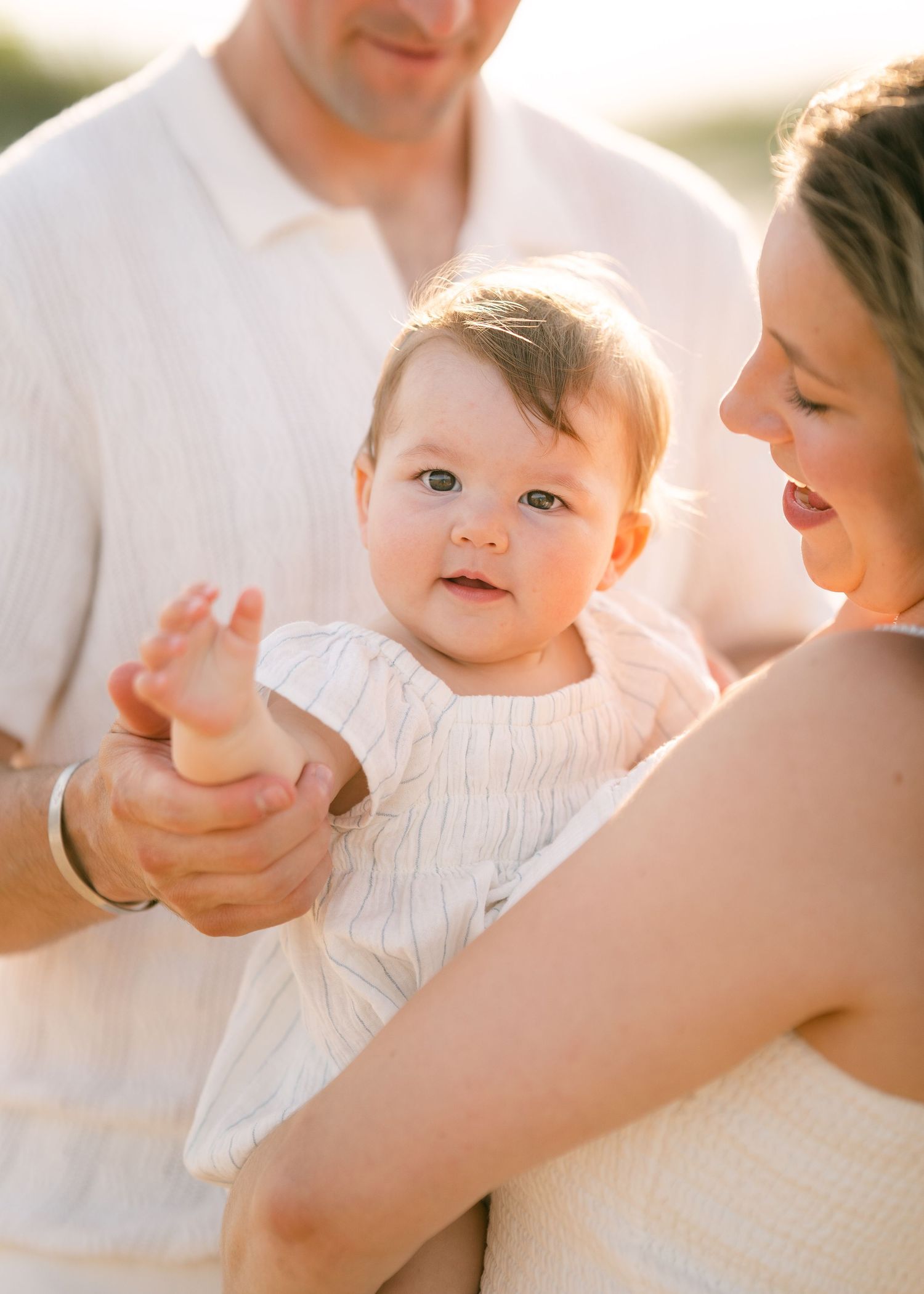 Close-up of adorable baby in white outfit being held by parents on sunny beach.