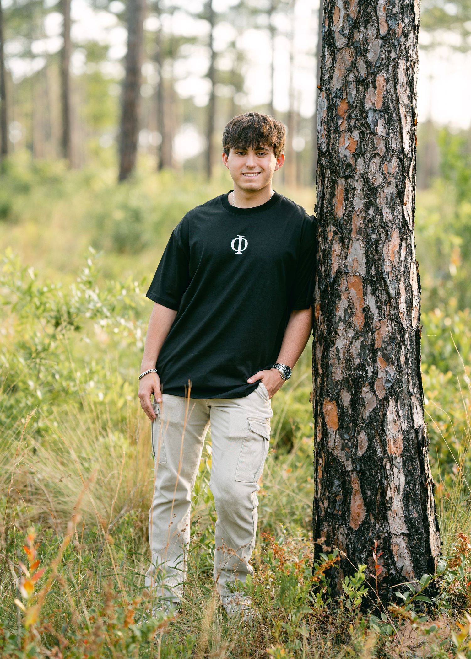 Teen boy in black t-shirt leaning against pine tree in natural forest setting for senior portrait.