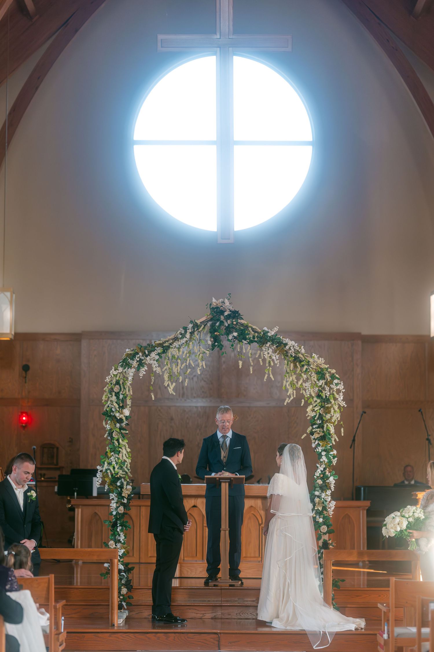 Bride and groom exchange vows beneath floral arch in church with stunning circular stained glass window.