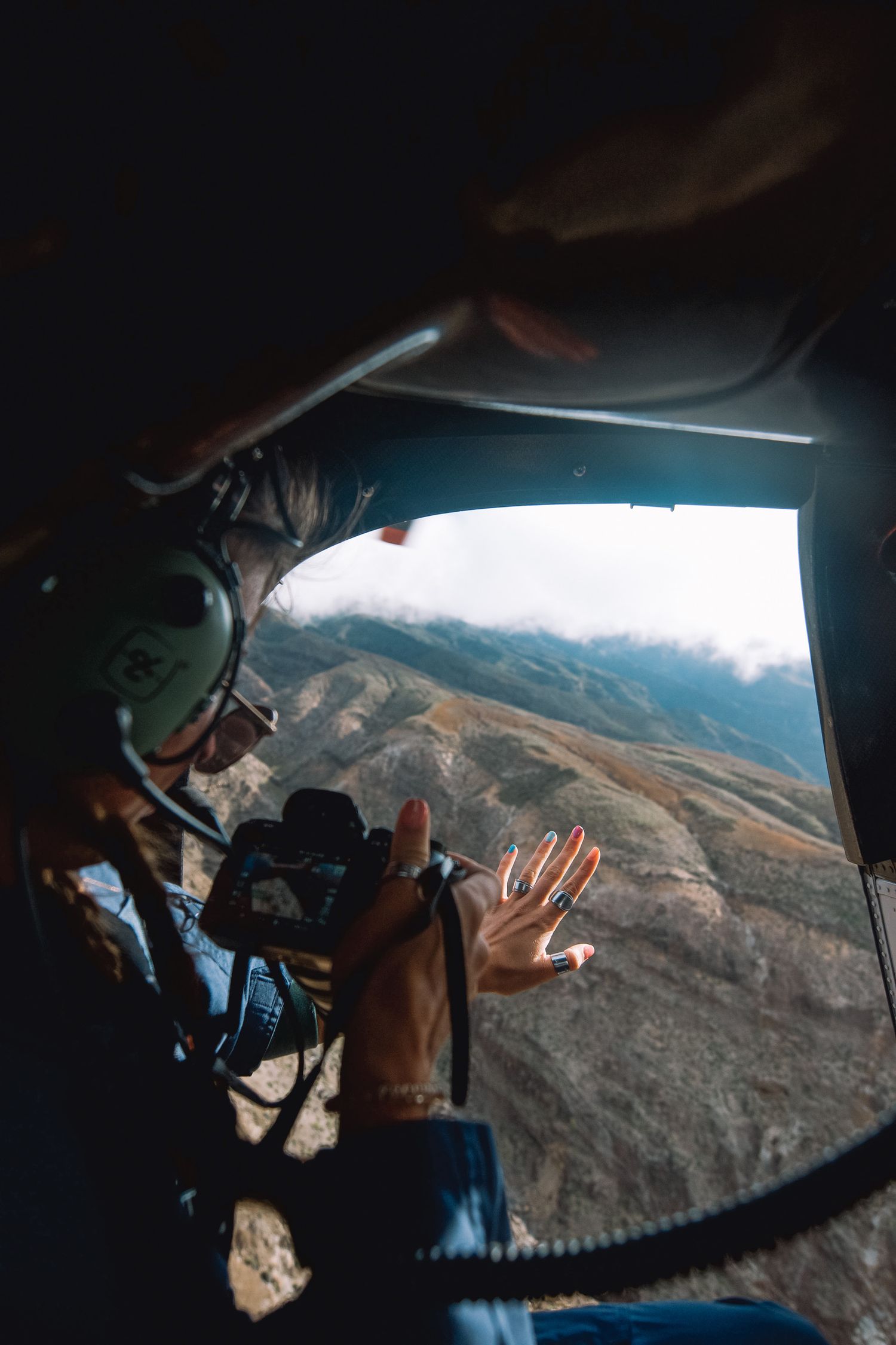 Photographer captures mountain landscape through open helicopter door wearing aviation headset mid-flight.