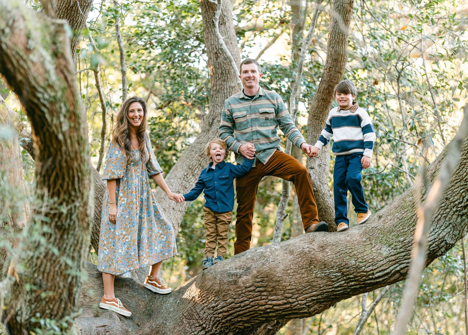 Happy family of four posing on large oak tree branch in forest setting.