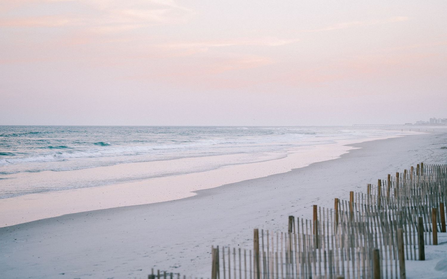 Peaceful pink and lavender sunrise over calm ocean waves with white sandy beach and wooden sand fence.