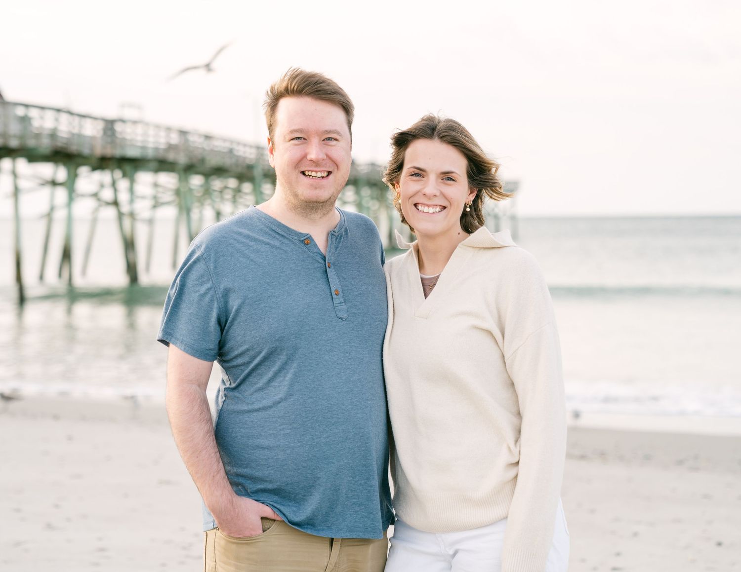 Smiling couple standing together on beach with pier behind them, dressed casually in blue and cream.