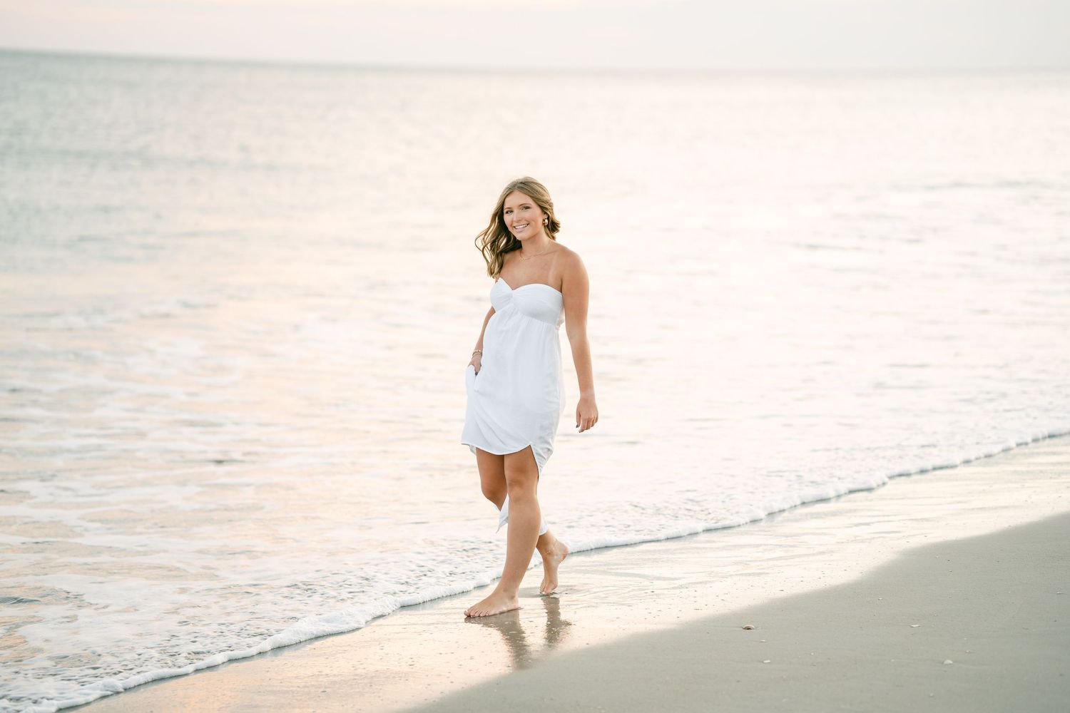 Young woman in white strapless dress walking barefoot along a serene beach at sunset.